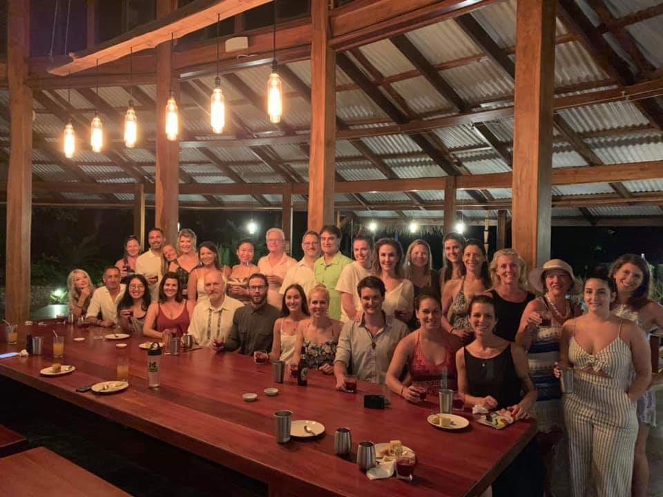 Roughly thirty smiling retreat participants raise glasses for a toast behind a long wooden table in an open-air pavilion at night, lit by warm pendant bulbs.