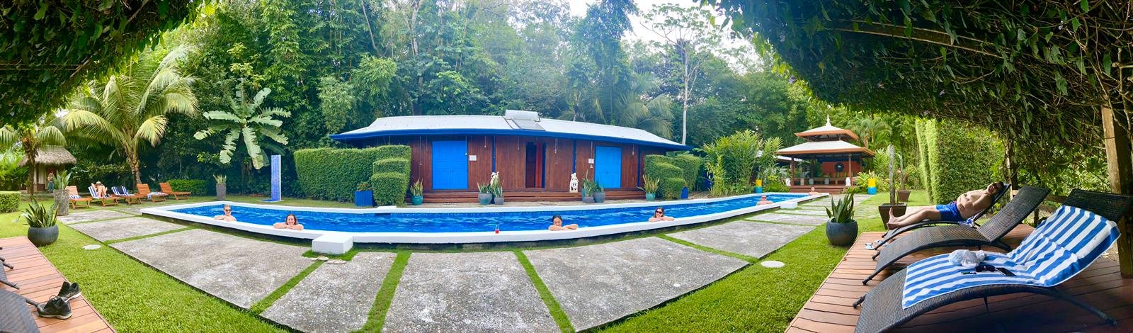 Wide panoramic view of the bright blue lap pool at Blue Osa with four women relaxing in the water, lounge chairs on the patio, wooden buildings with turquoise doors, and lush jungle surrounding the resort.