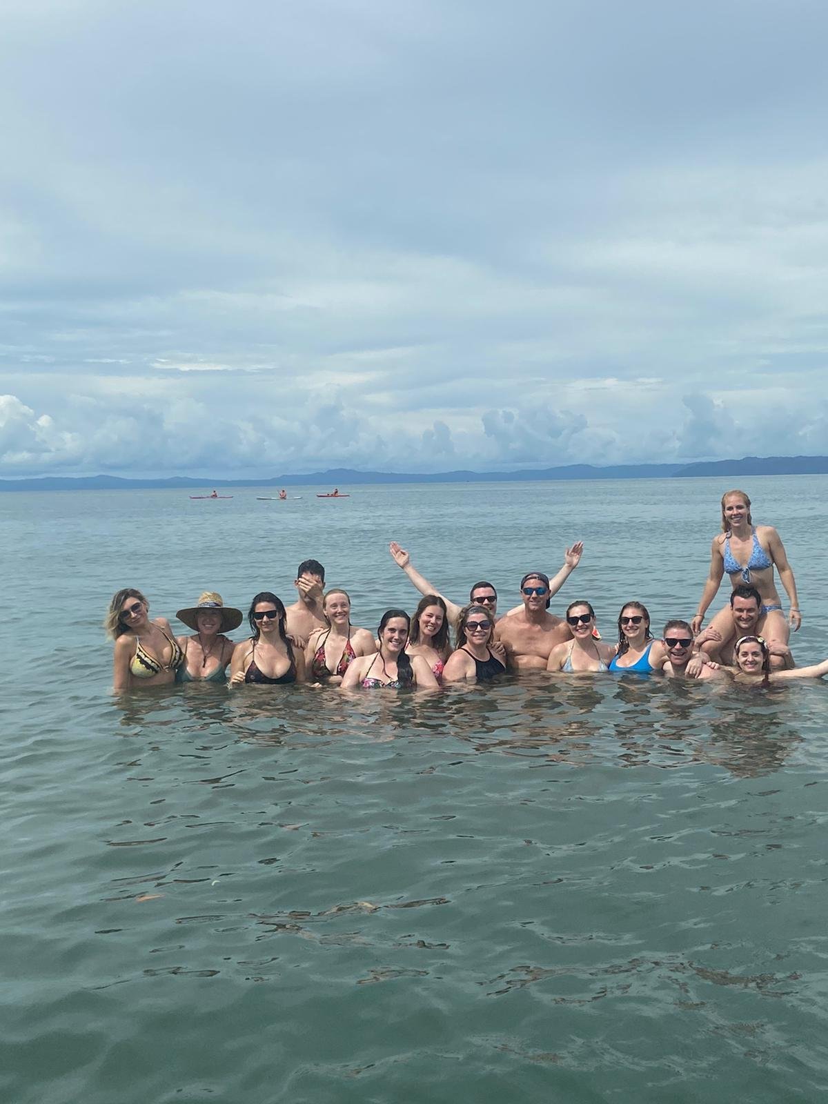 About fifteen retreat participants smile waist-deep in calm ocean water during a group swim, with one woman sitting on a man's shoulders, distant hills on the horizon, and soft cloudy skies.
