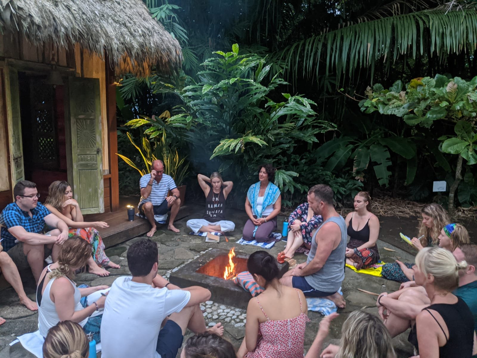 Roughly fifteen retreat participants sit in a circle on stone pavers around a small fire pit at Blue Osa, surrounded by jungle greenery and a thatched-roof building during an evening sharing circle.