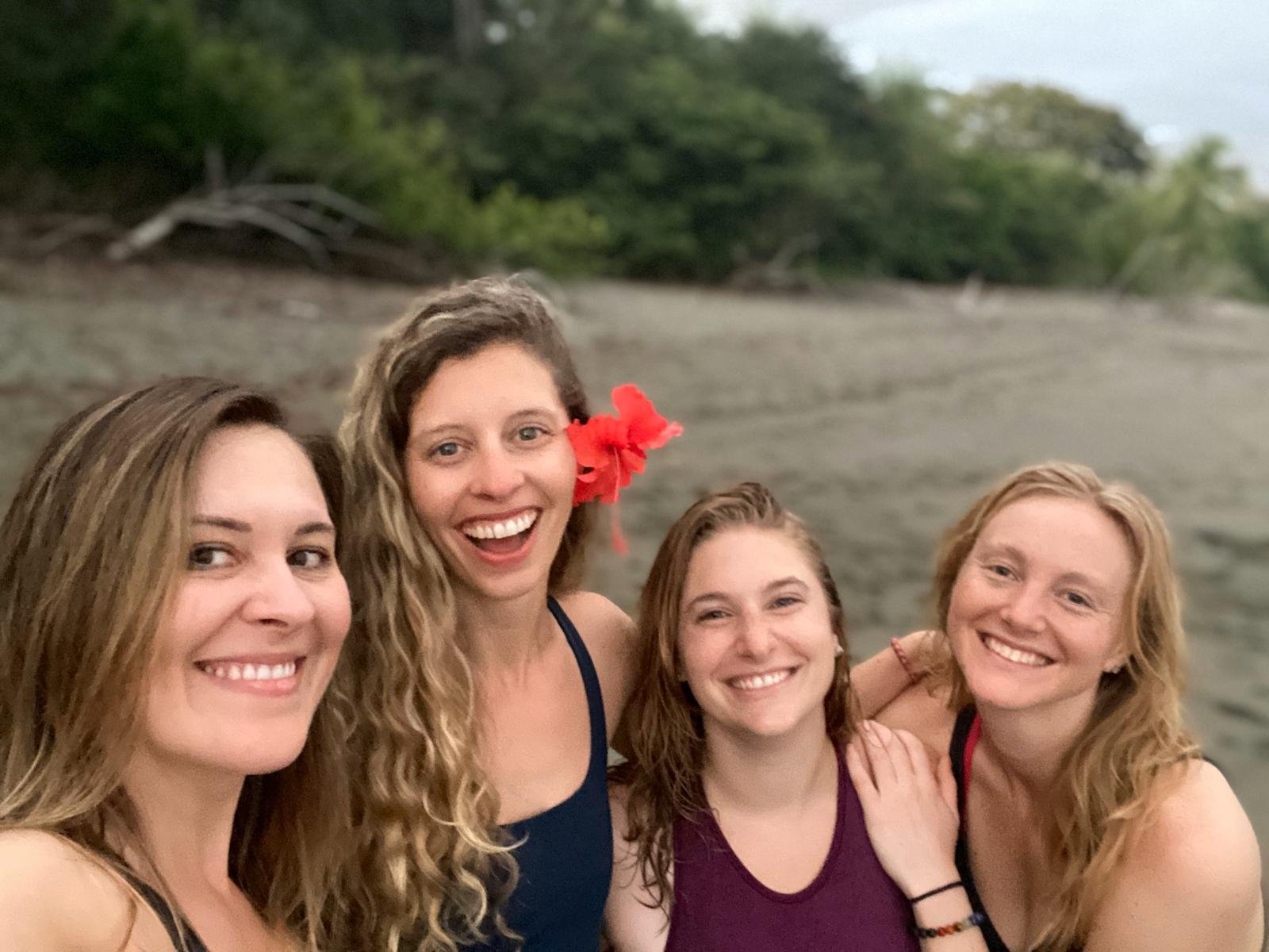 Four women smile for a beach selfie on dark volcanic sand in Costa Rica, one with a red hibiscus behind her ear, with a green jungle treeline and overcast sky behind them.