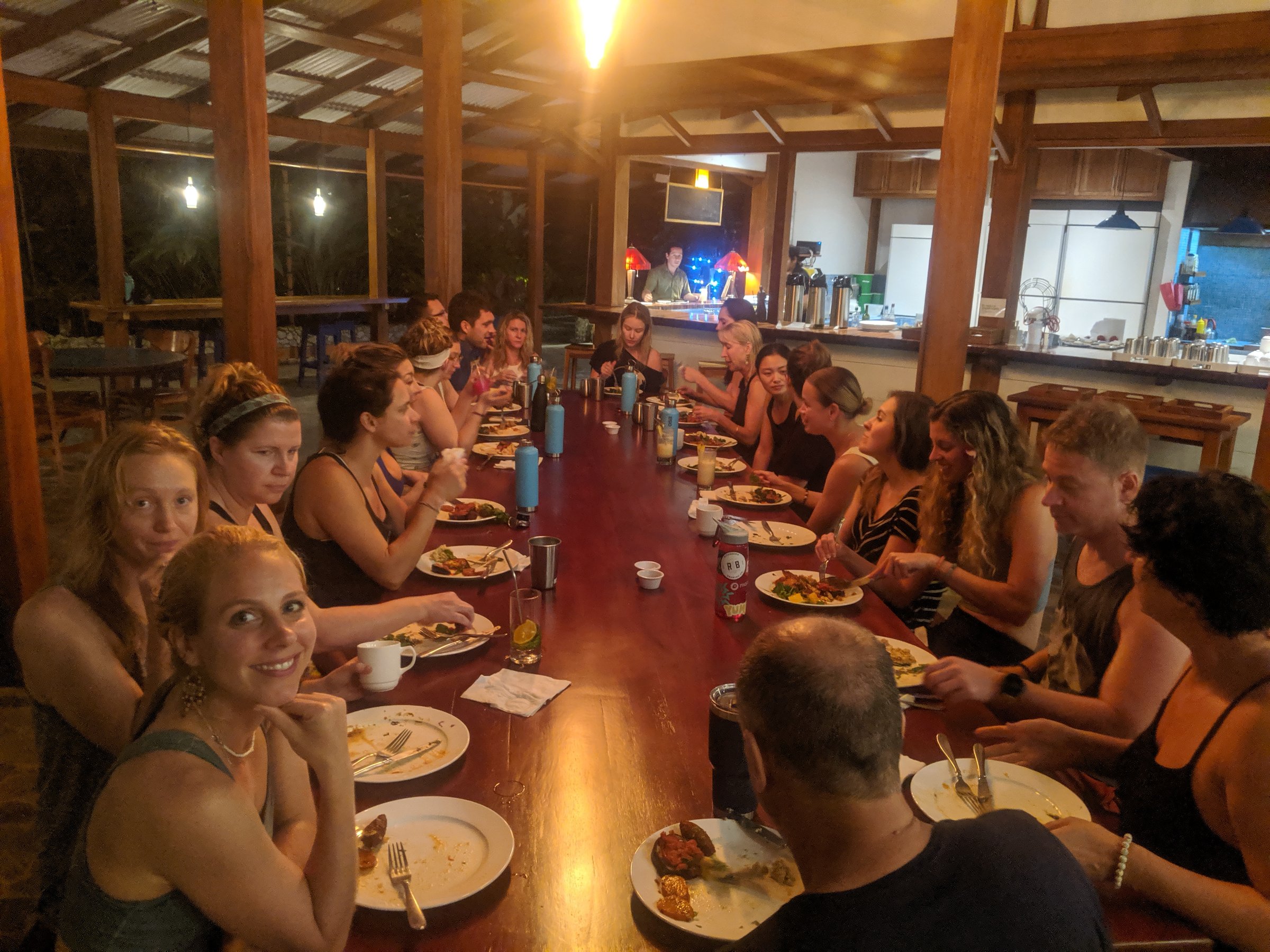 About twenty retreat participants share a communal dinner at a long wooden table in an open-air pavilion at Blue Osa at night, with plates, drinks, and warm lighting as people talk and eat.