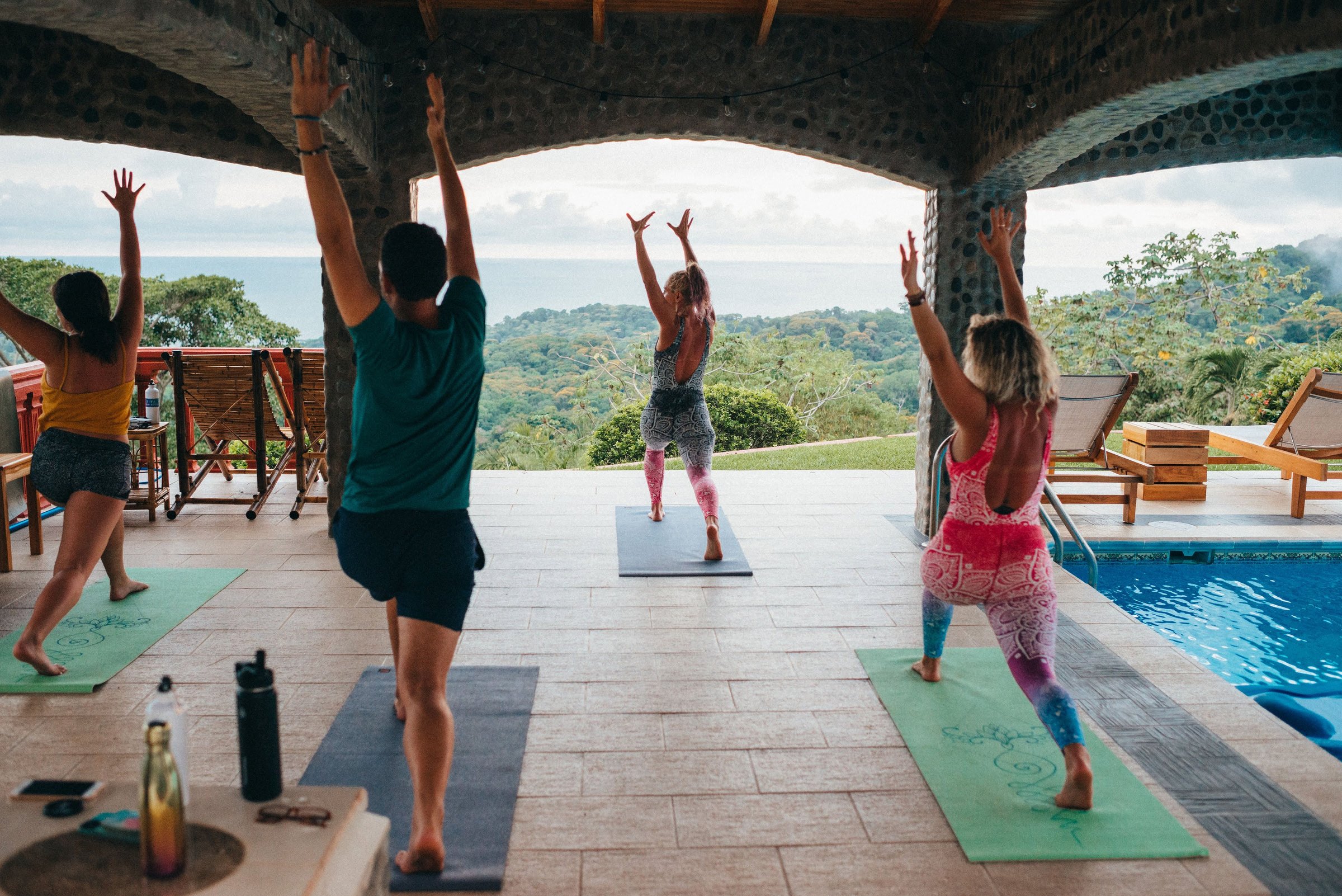 Four people practice yoga with arms raised on mats under a stone-arched pavilion overlooking jungle hills, the ocean, and a pool to the right.