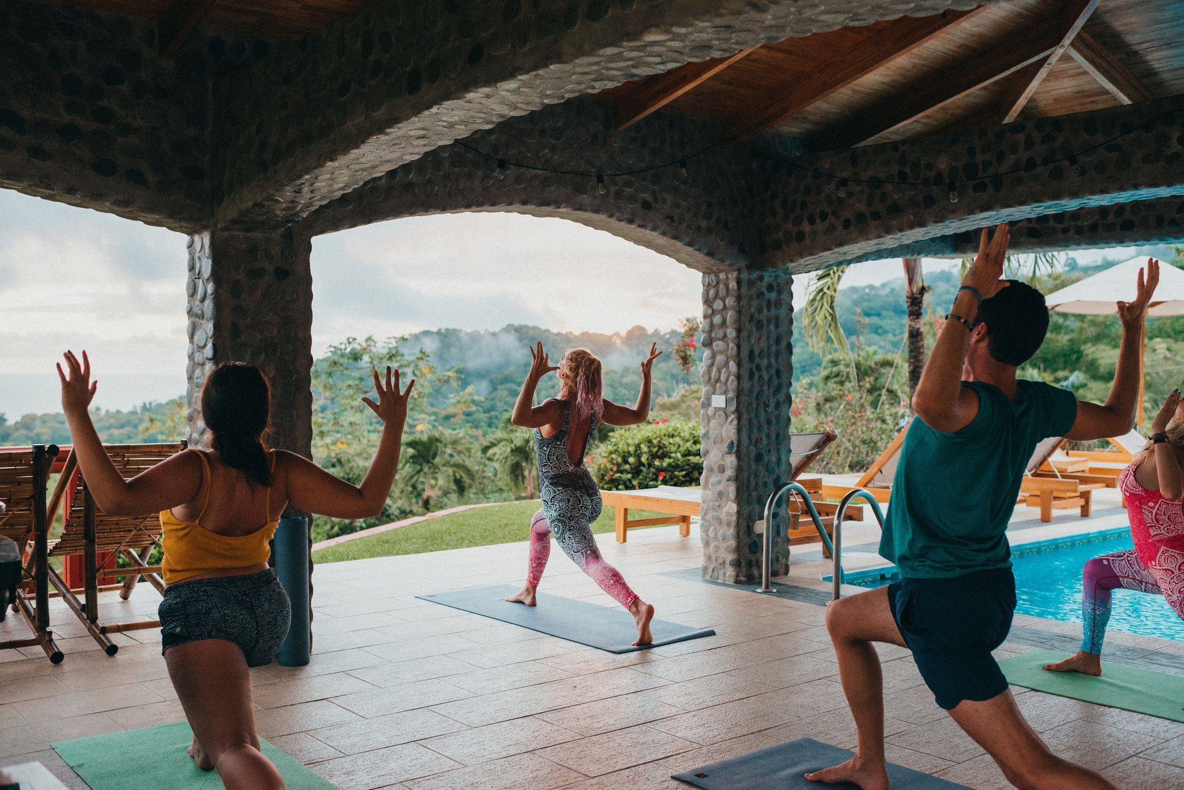 Retreat guests hold warrior poses on yoga mats beneath stone arches framing green hills and soft evening light beside a turquoise pool.