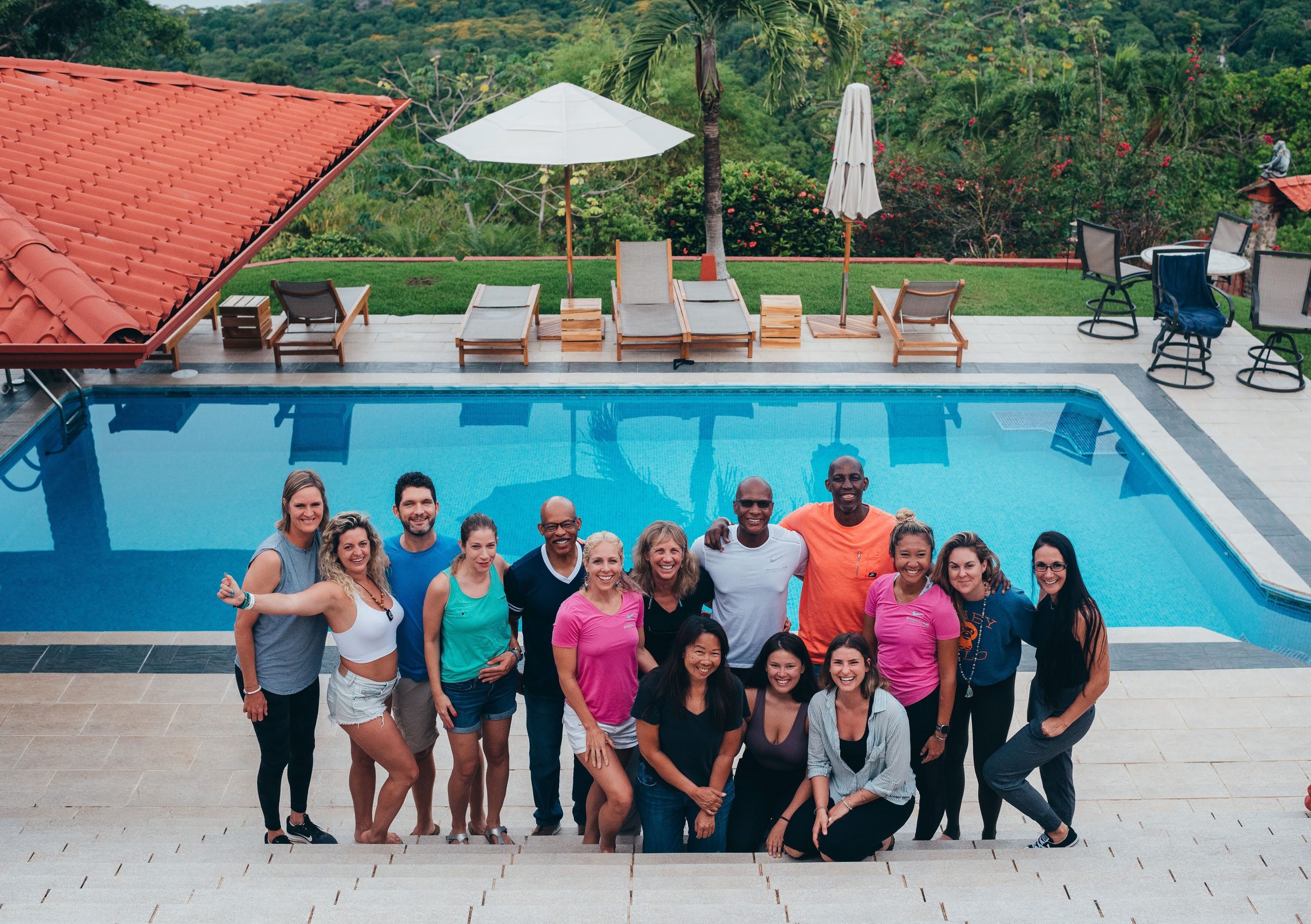 Fifteen retreat participants pose on a tiled patio beside a bright blue pool with green hills and tropical forest in the background.