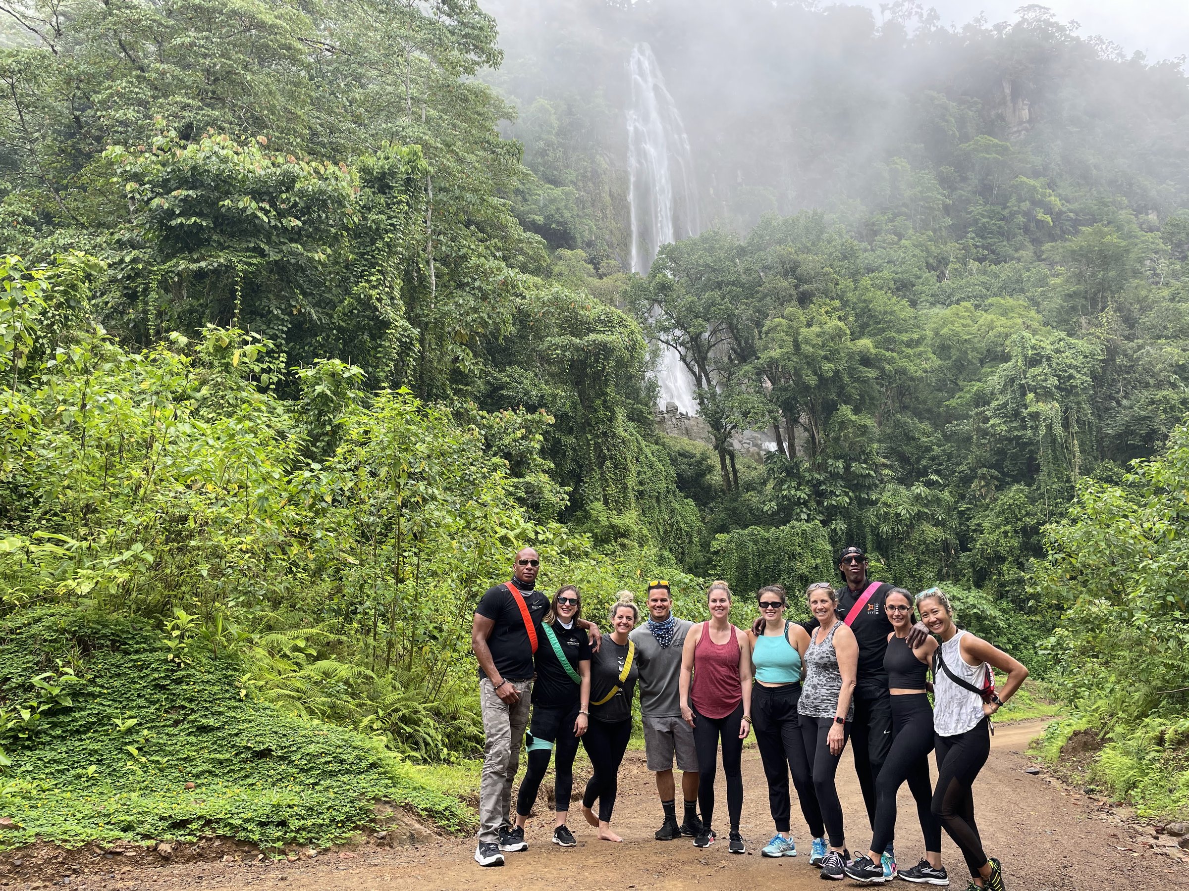 Ten retreat travelers stand on a dirt road in lush jungle with a tall waterfall cascading down a misty green mountainside behind them.