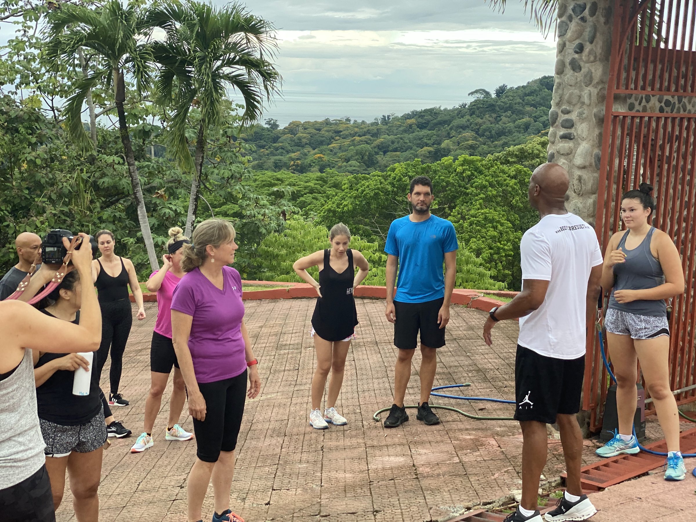 A group gathers on a circular stone terrace overlooking dense green jungle and the distant Pacific while a leader in a white shirt addresses them.