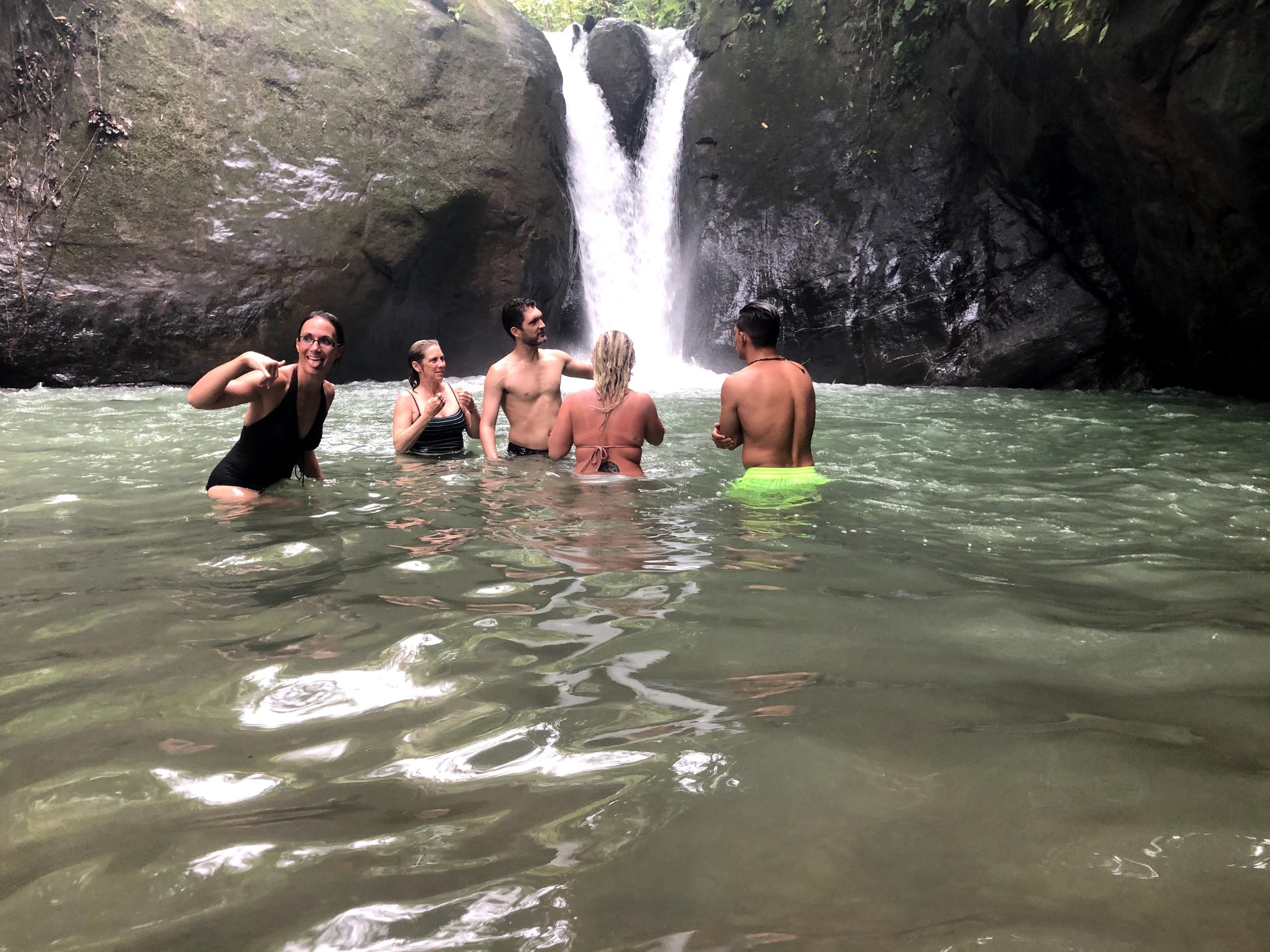Five retreat guests swim chest-deep in a greenish natural pool at the base of a narrow waterfall between mossy rock walls.