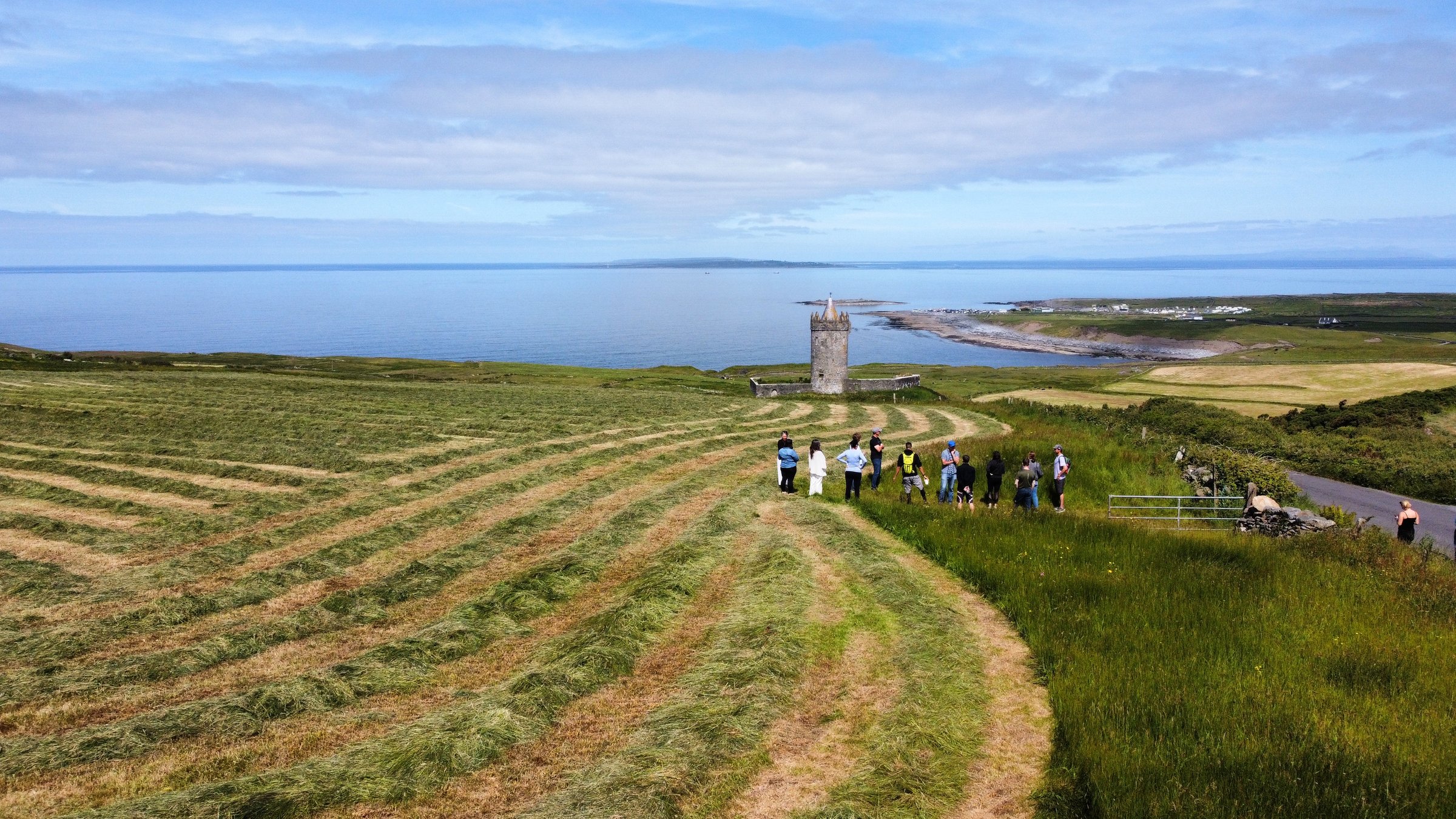 A group stands in a hay-lined green field overlooking Doonagore Castle and the Atlantic Ocean.
