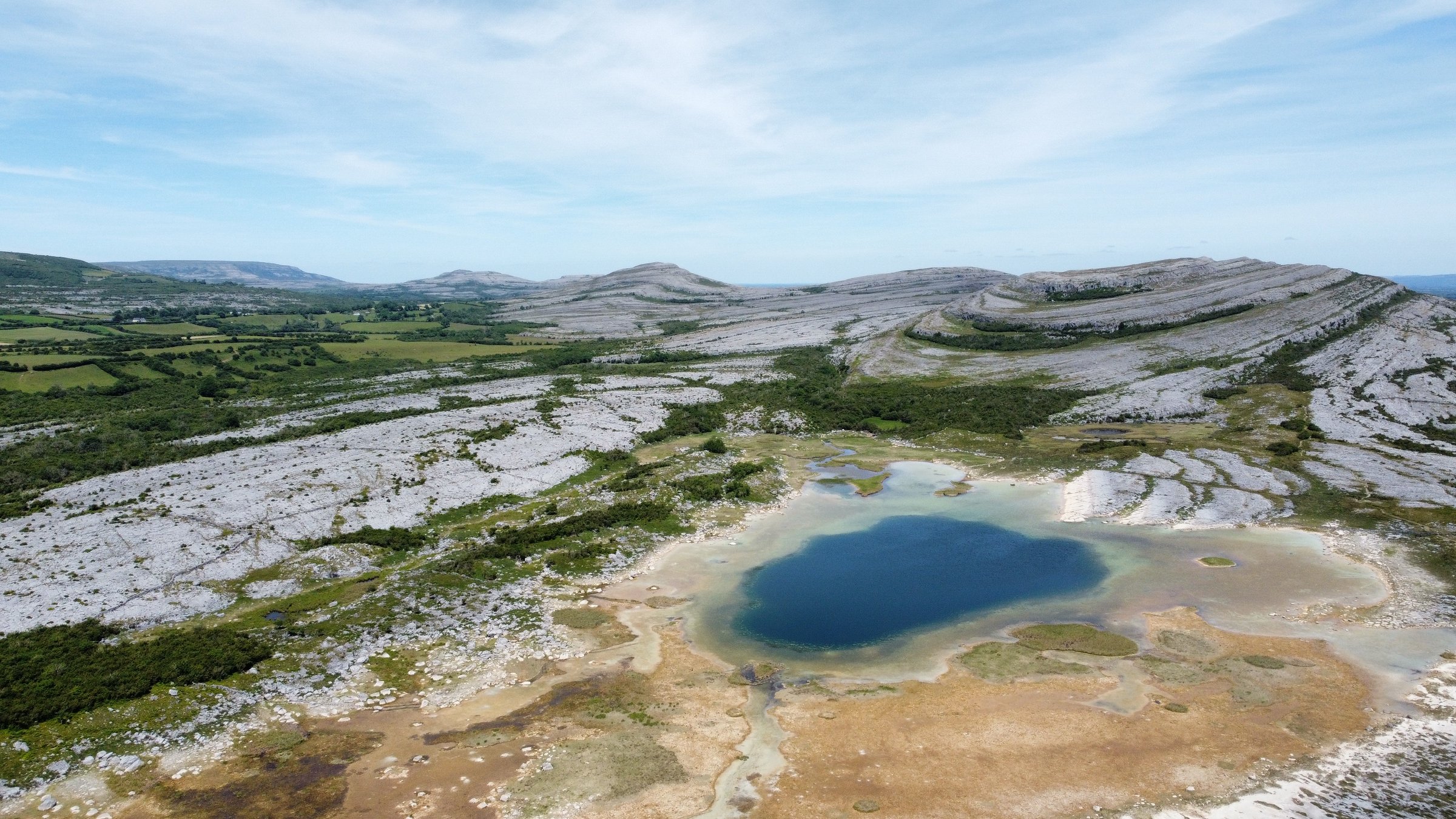 Aerial view of the Burren's limestone karst landscape with a bright blue lake and rocky hills.