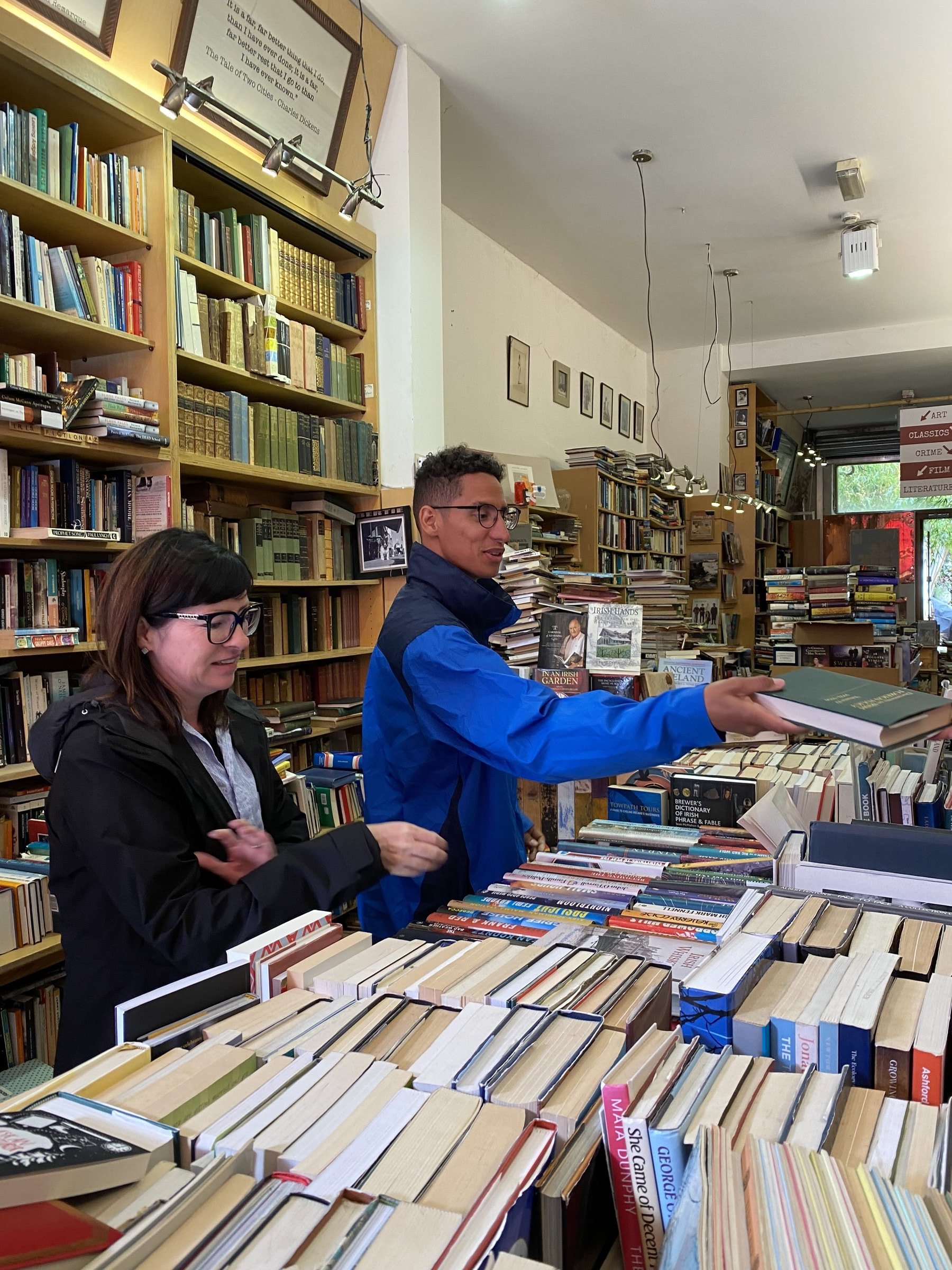 Two people browse through stacks and shelves of books in a cozy, crowded bookstore.