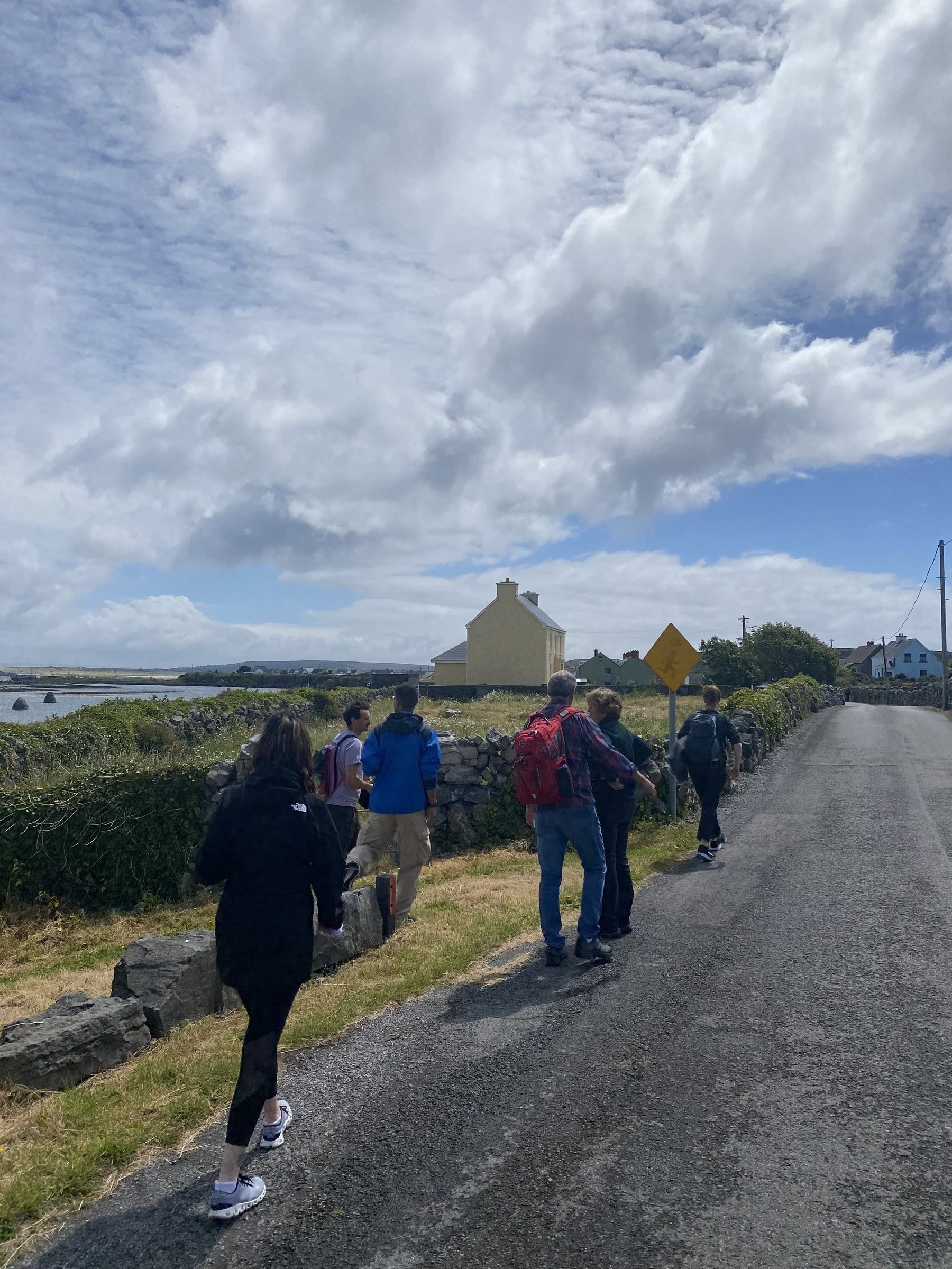 A group walks along a narrow coastal road past a stone wall toward a yellow house and the sea.