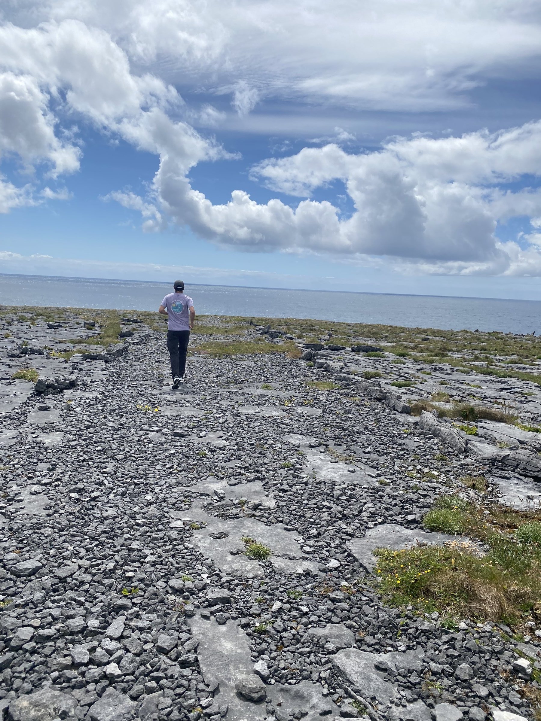 A person in a purple shirt hikes across the rugged limestone landscape of the Burren toward the ocean.