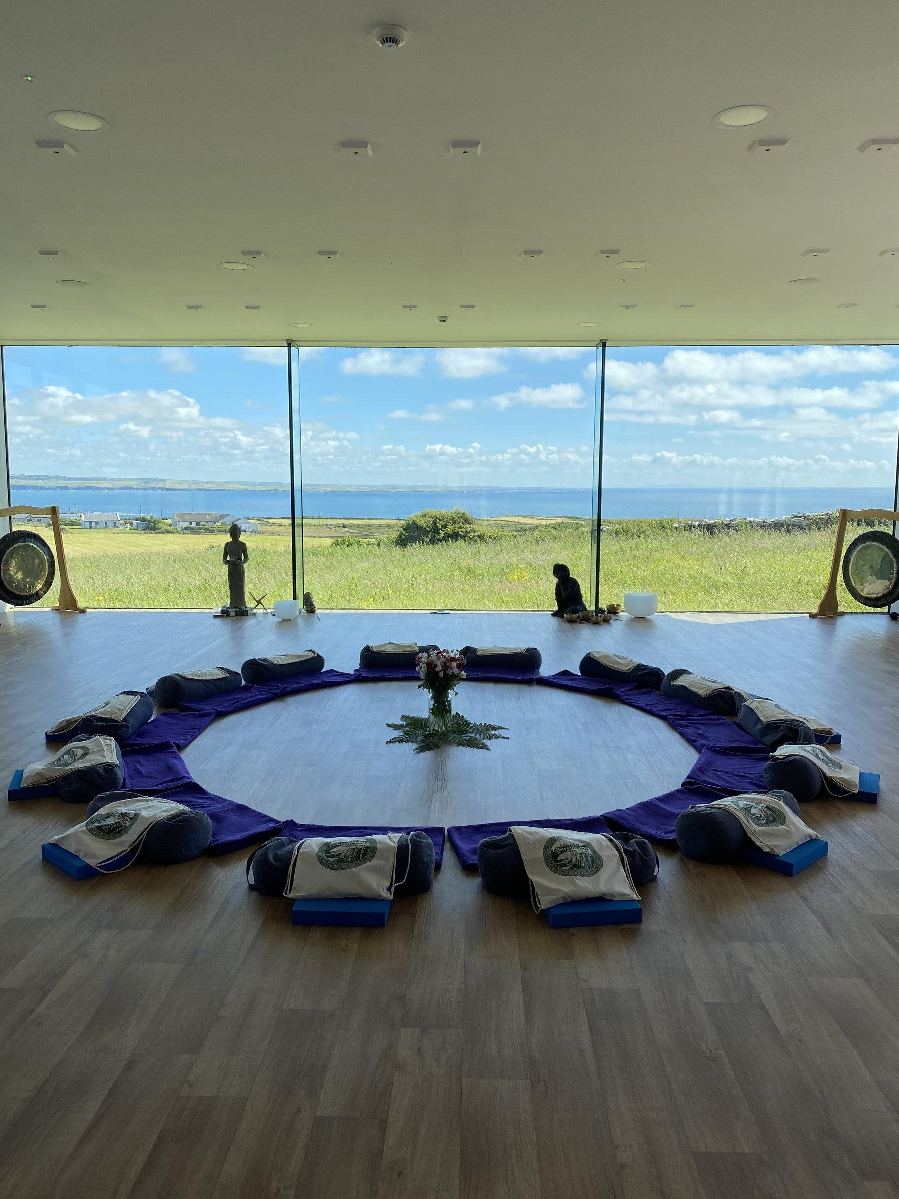 A circular arrangement of purple meditation mats and cushions fills a bright studio overlooking green fields and the sea.