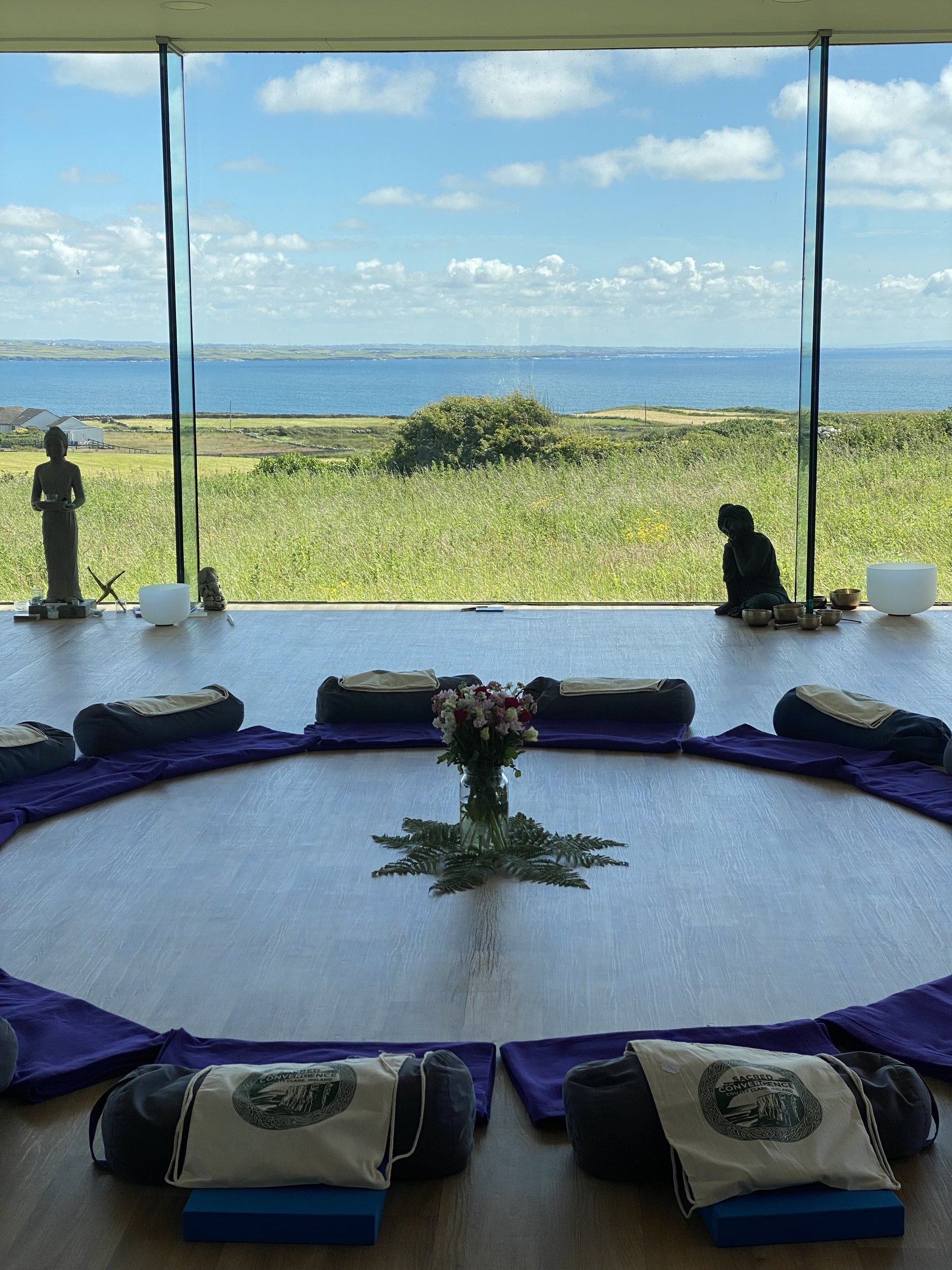 Sacred Convergence tote bags and meditation cushions circle a floral centerpiece in a coastal studio.