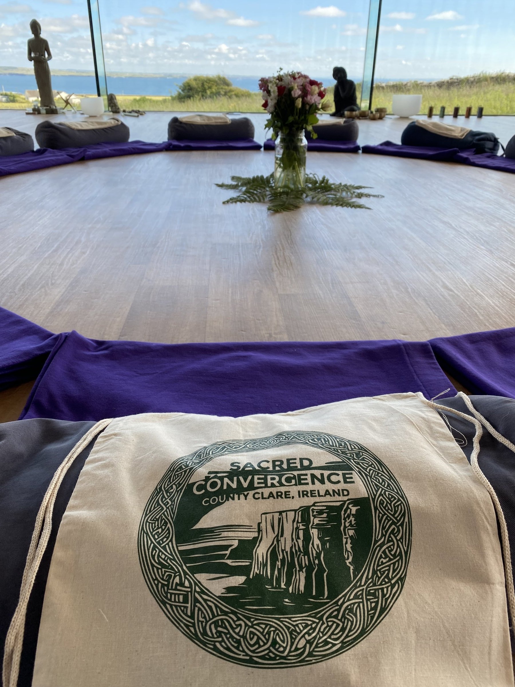 A Sacred Convergence canvas tote bag rests on purple fabric in a meditation studio with an ocean view.