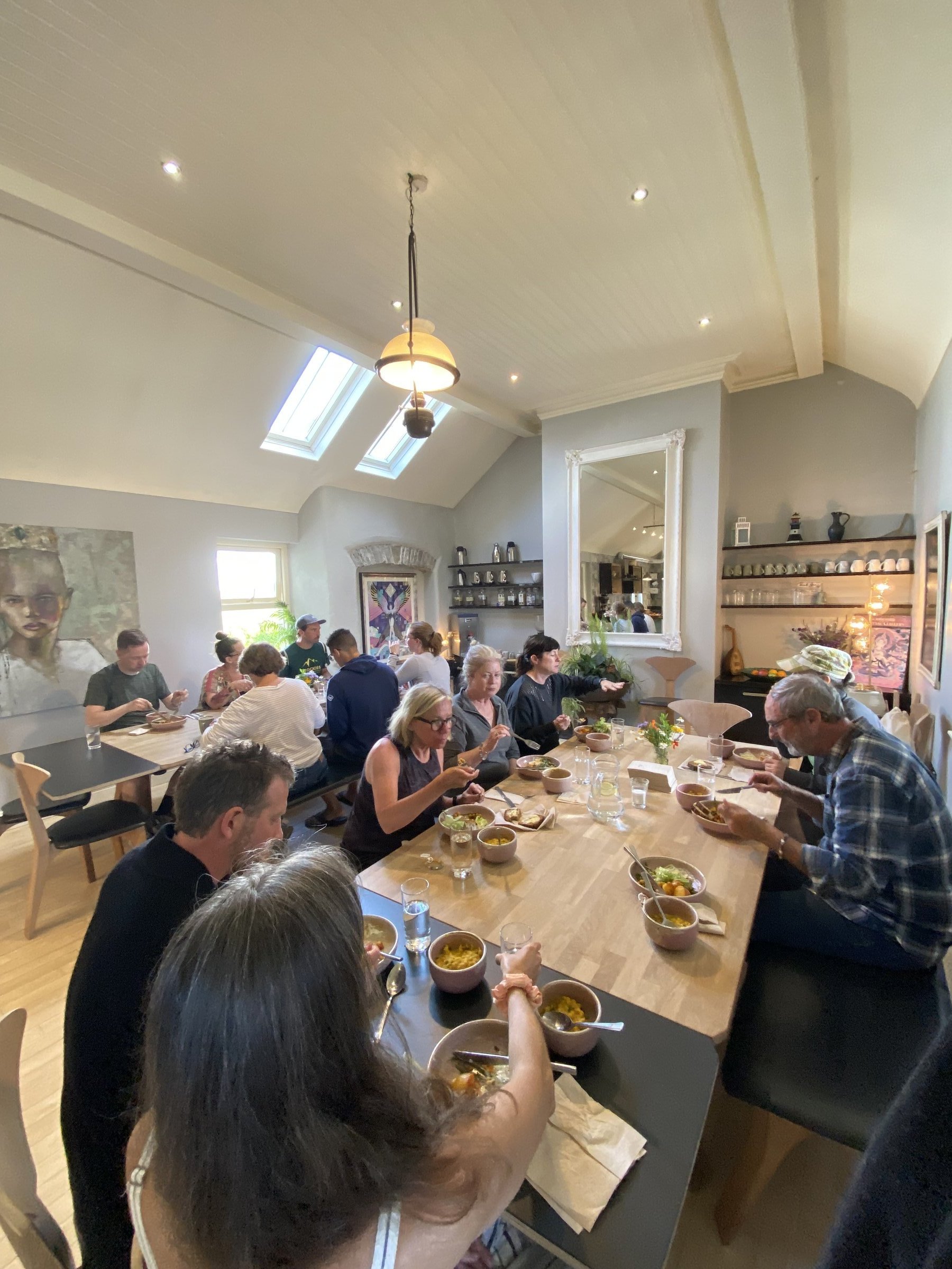 A group shares a communal meal around a long wooden table in a bright dining room.