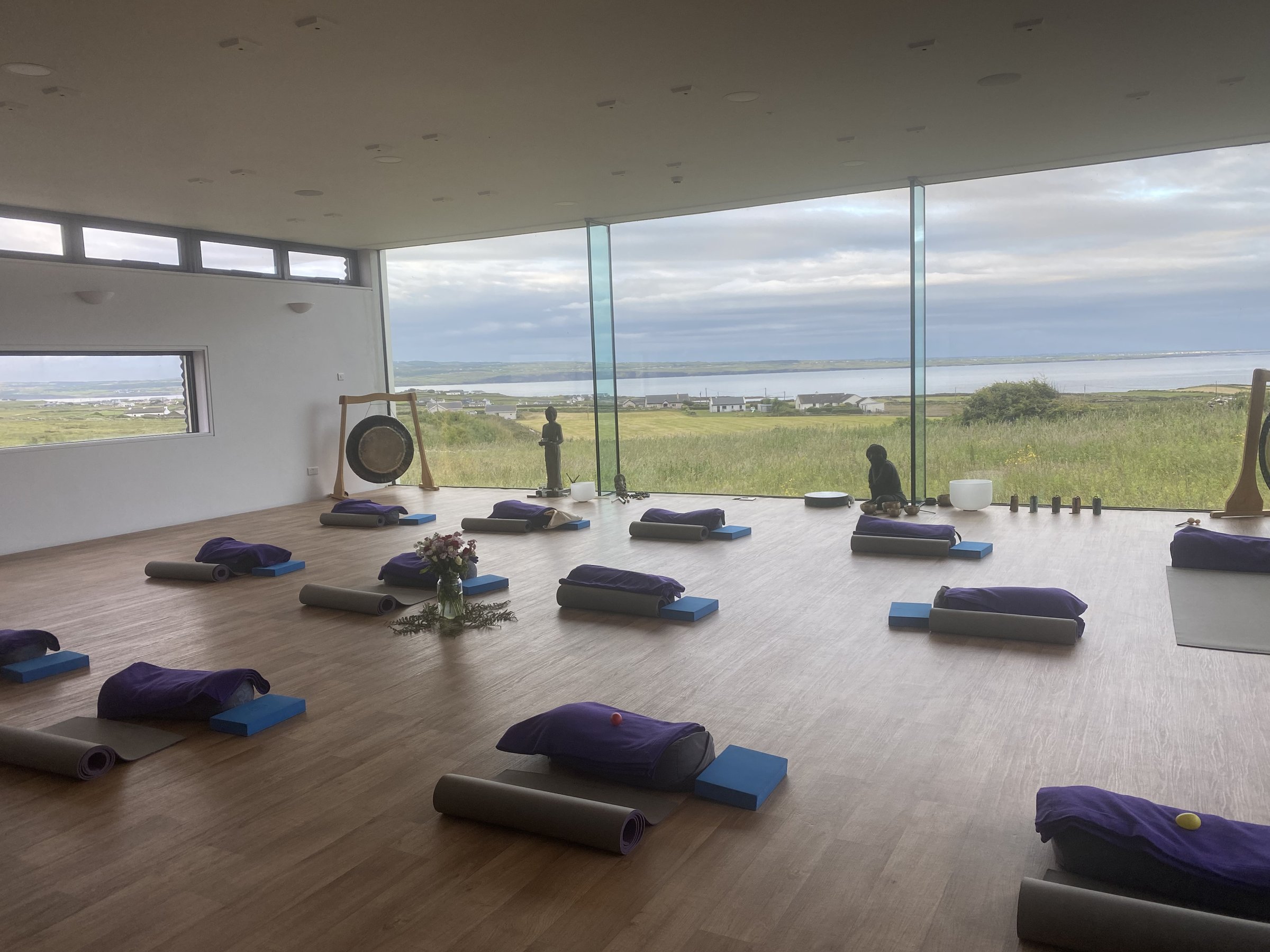 Yoga mats, purple blankets, and gongs are laid out in a studio with floor-to-ceiling coastal windows.
