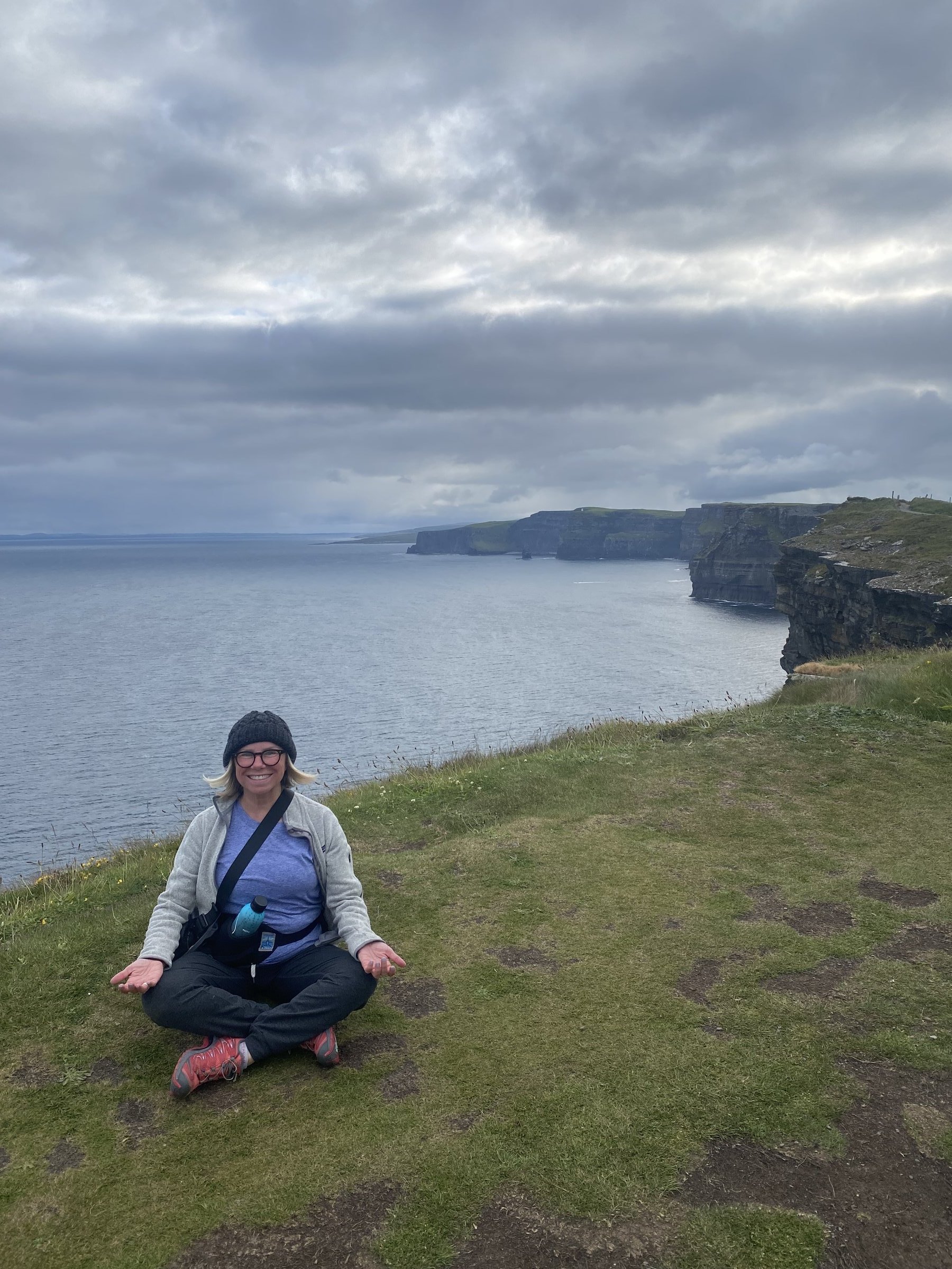 A woman sits cross-legged in meditation on a grassy cliff overlooking the Cliffs of Moher and Atlantic.