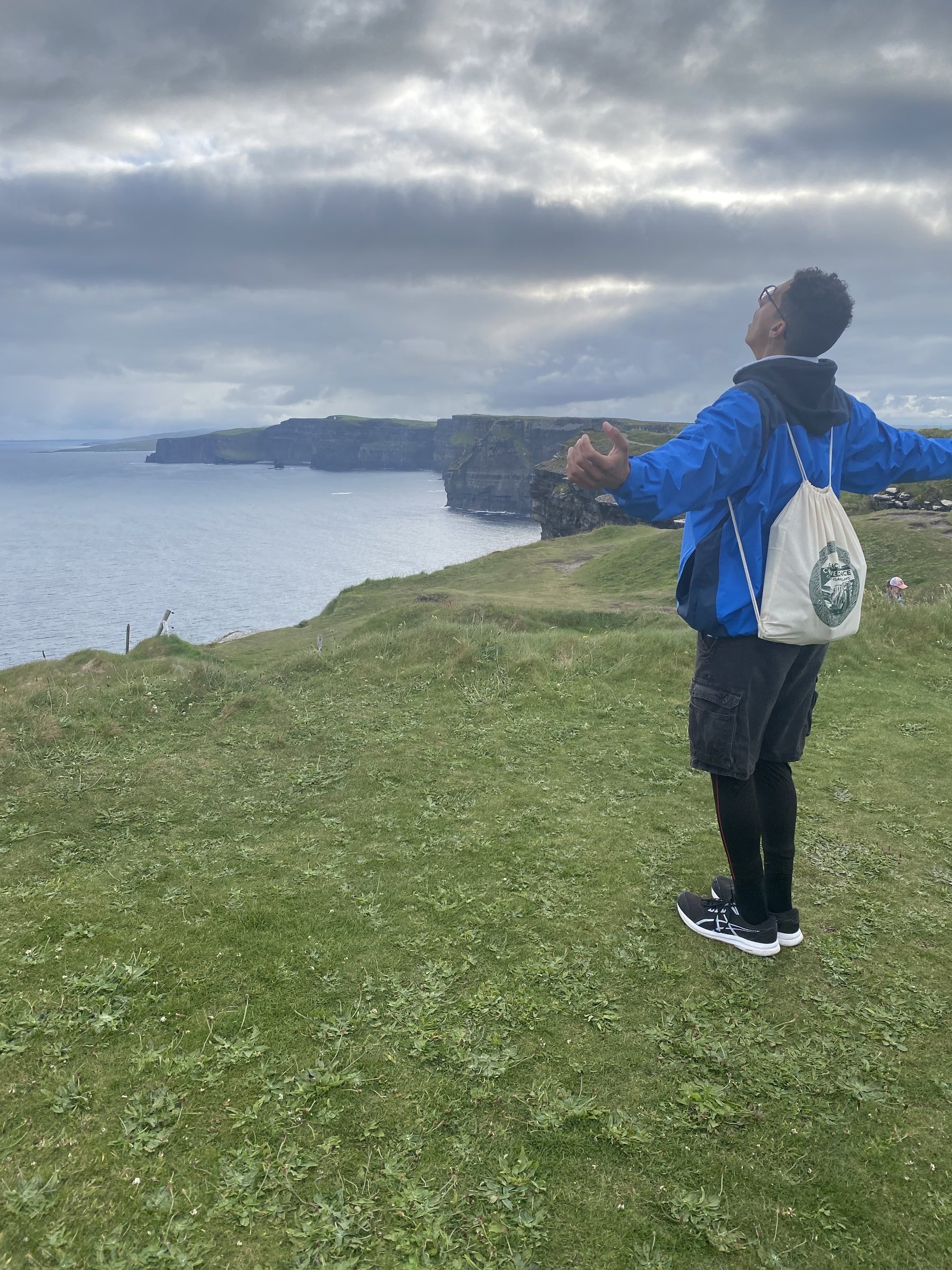 A hiker in a blue jacket stands on a grassy cliff edge overlooking the dramatic Cliffs of Moher and the Atlantic under heavy grey clouds.