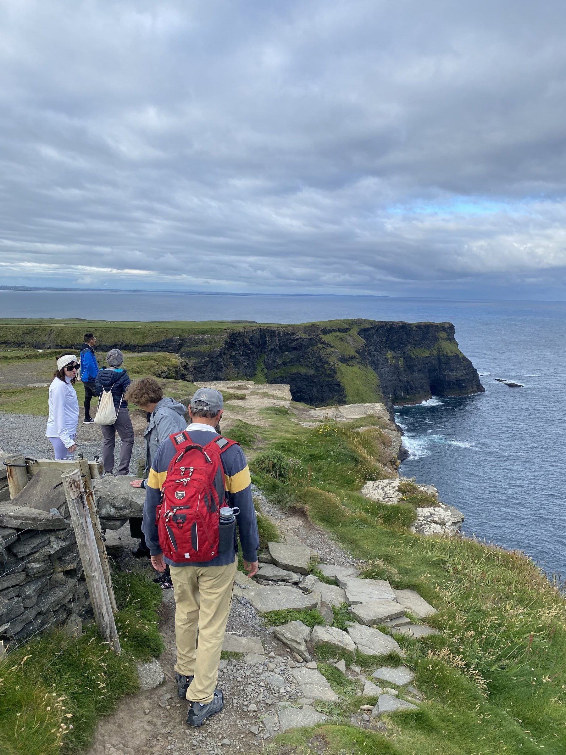Hikers walk along a rugged stone path beside the sheer Cliffs of Moher as waves crash against the dark rock below.