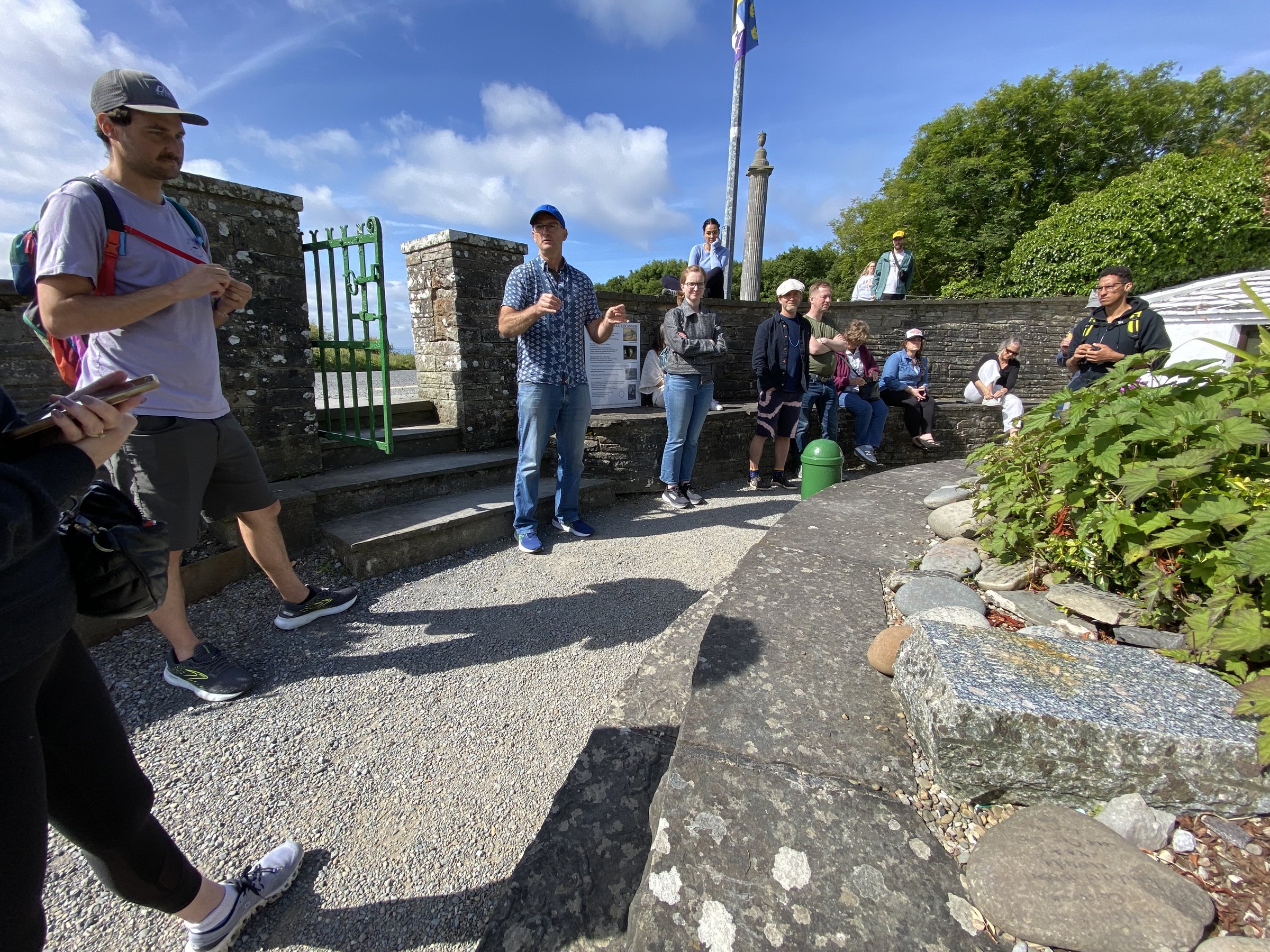 A tour group gathers in a sunny gravel courtyard with stone walls while a guide in a blue cap speaks and gestures.