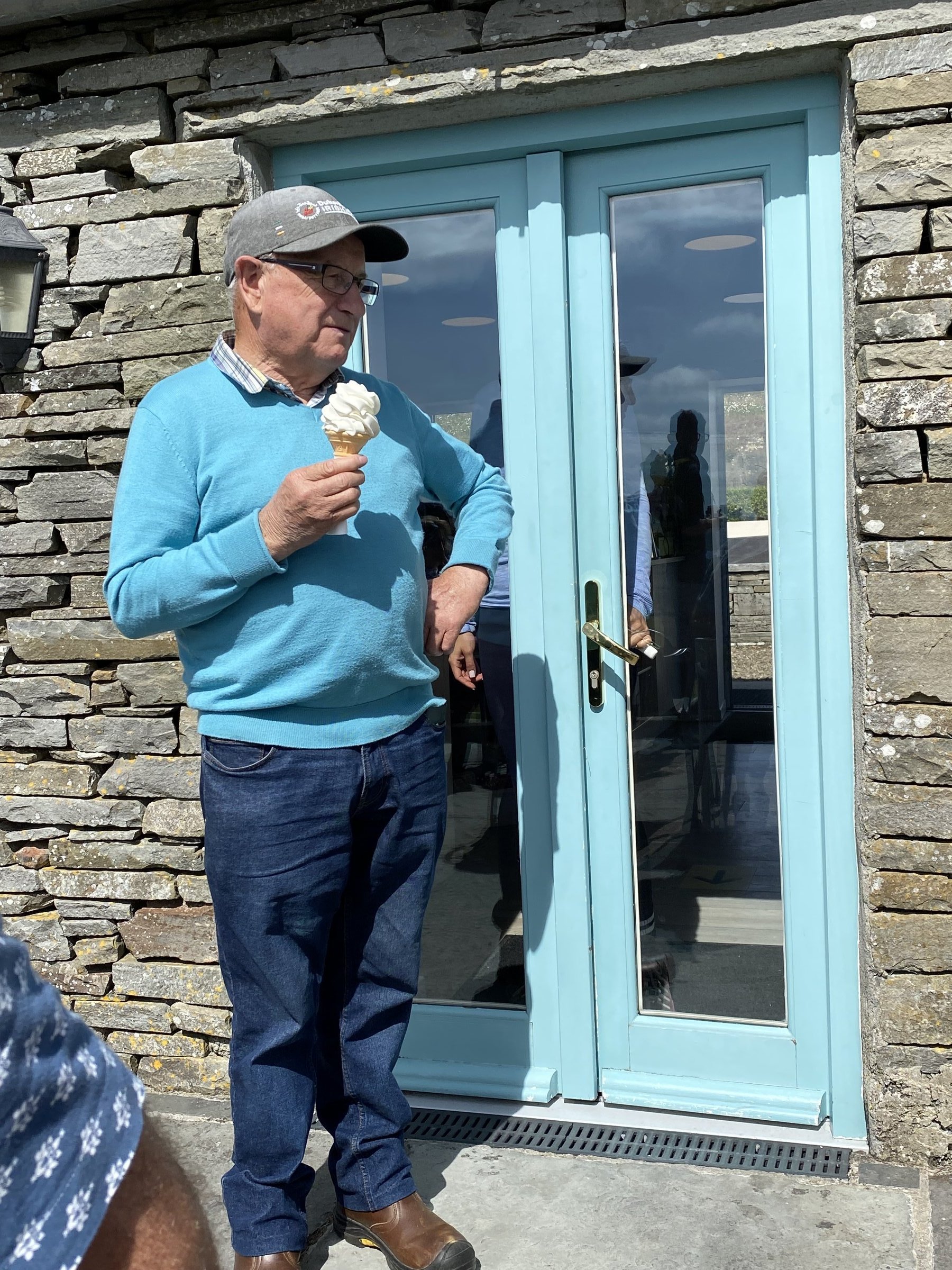 An older man in a grey cap and light blue sweater enjoys a soft-serve ice cream cone outside a stone building with pale blue doors.