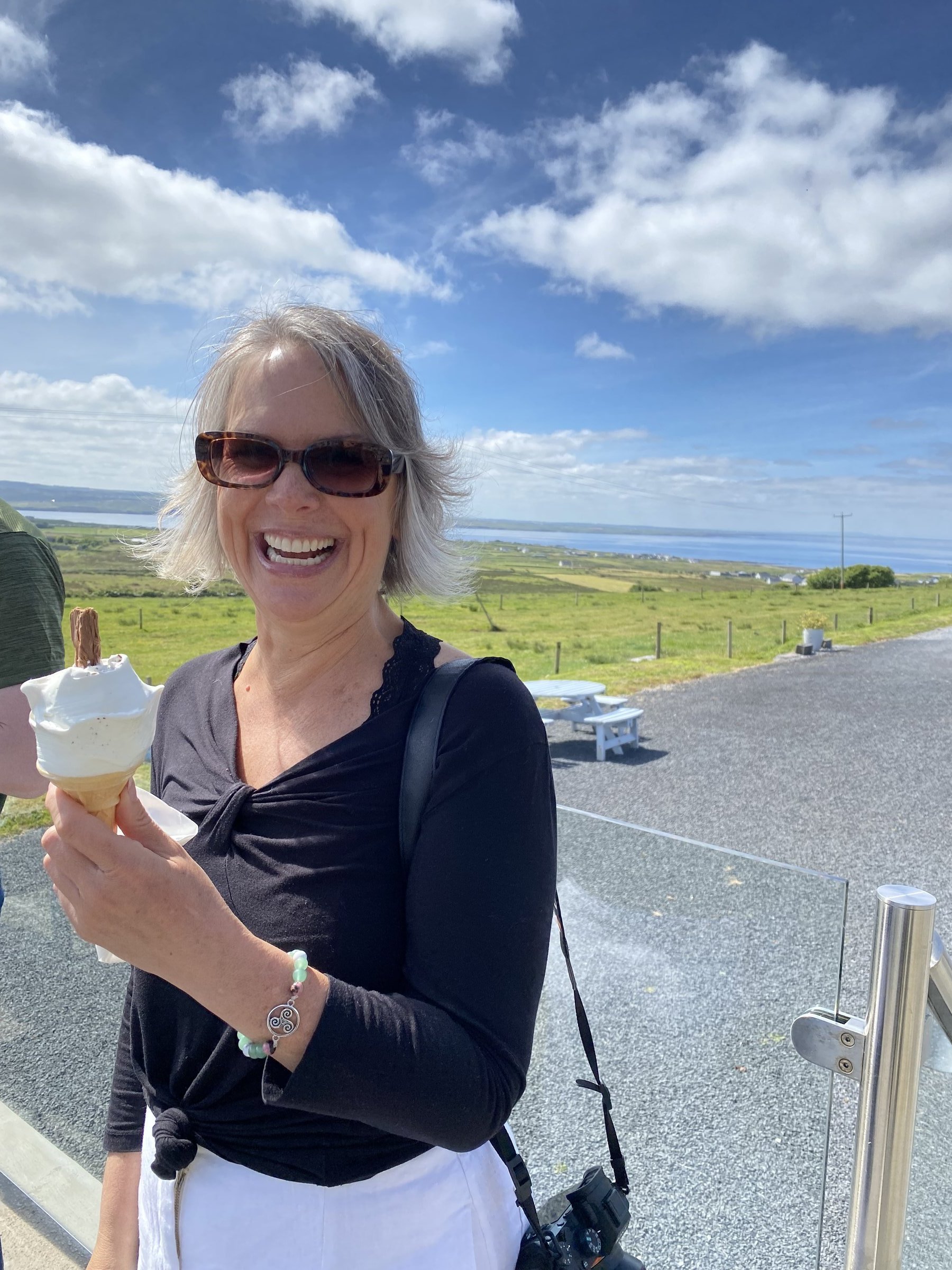 A smiling woman with sunglasses holds a soft-serve cone topped with a chocolate flake at a coastal viewpoint overlooking green fields and the sea.