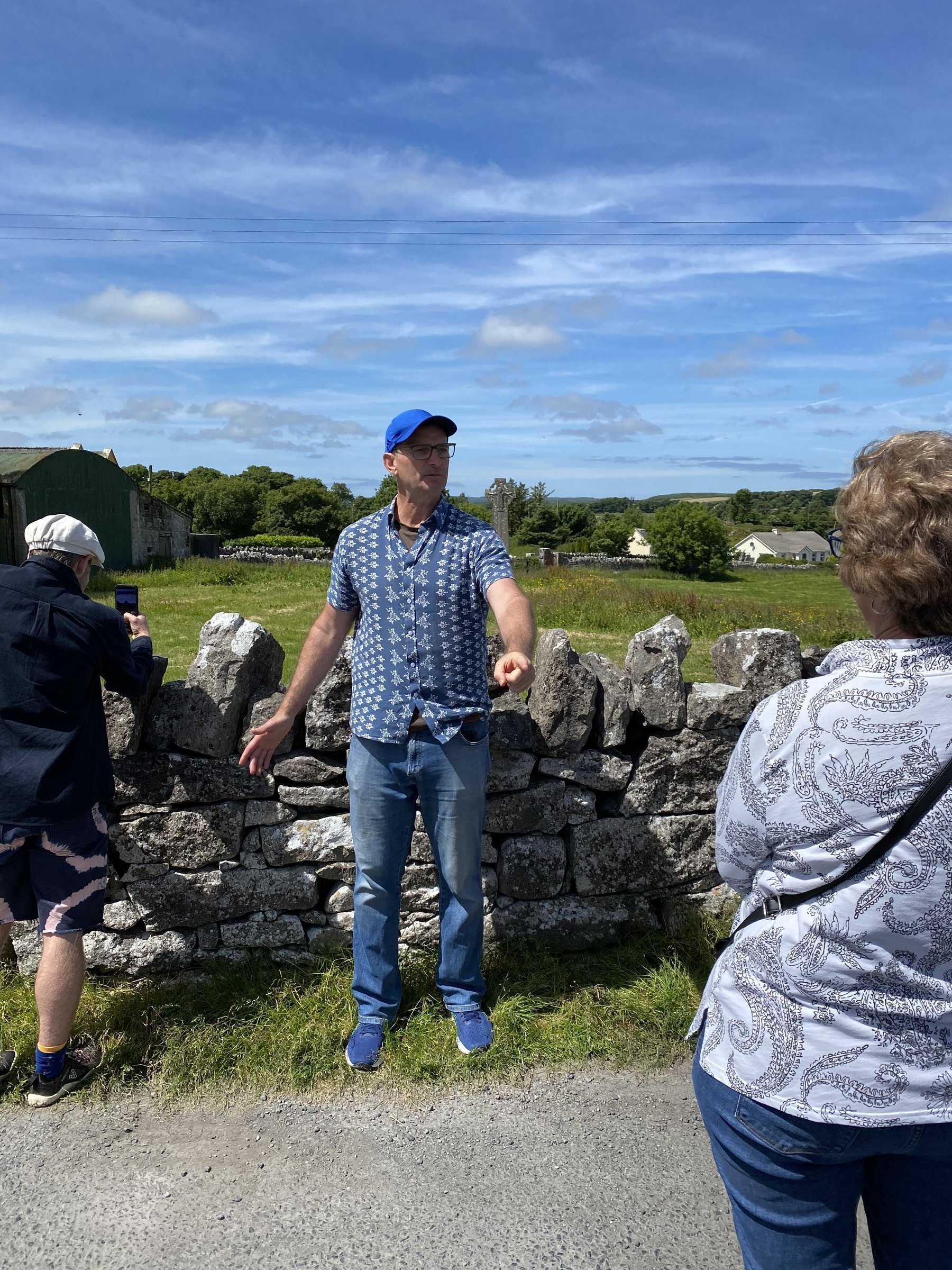 A guide in a blue patterned shirt and cap talks beside a traditional dry-stone wall in rolling green Irish countryside under a bright blue sky.