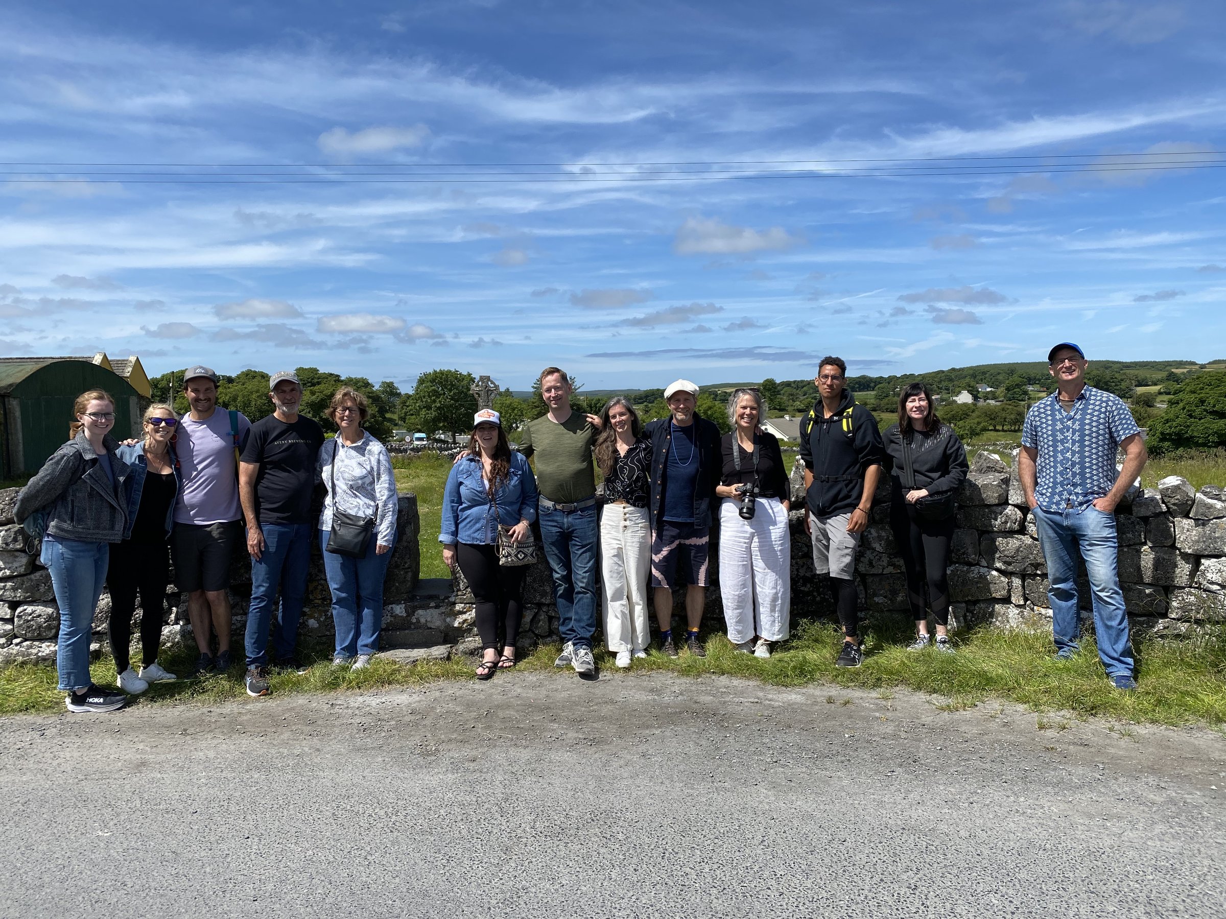 Thirteen retreat participants smile together for a group photo in front of a stone wall with green hills and a Celtic cross in the distance.