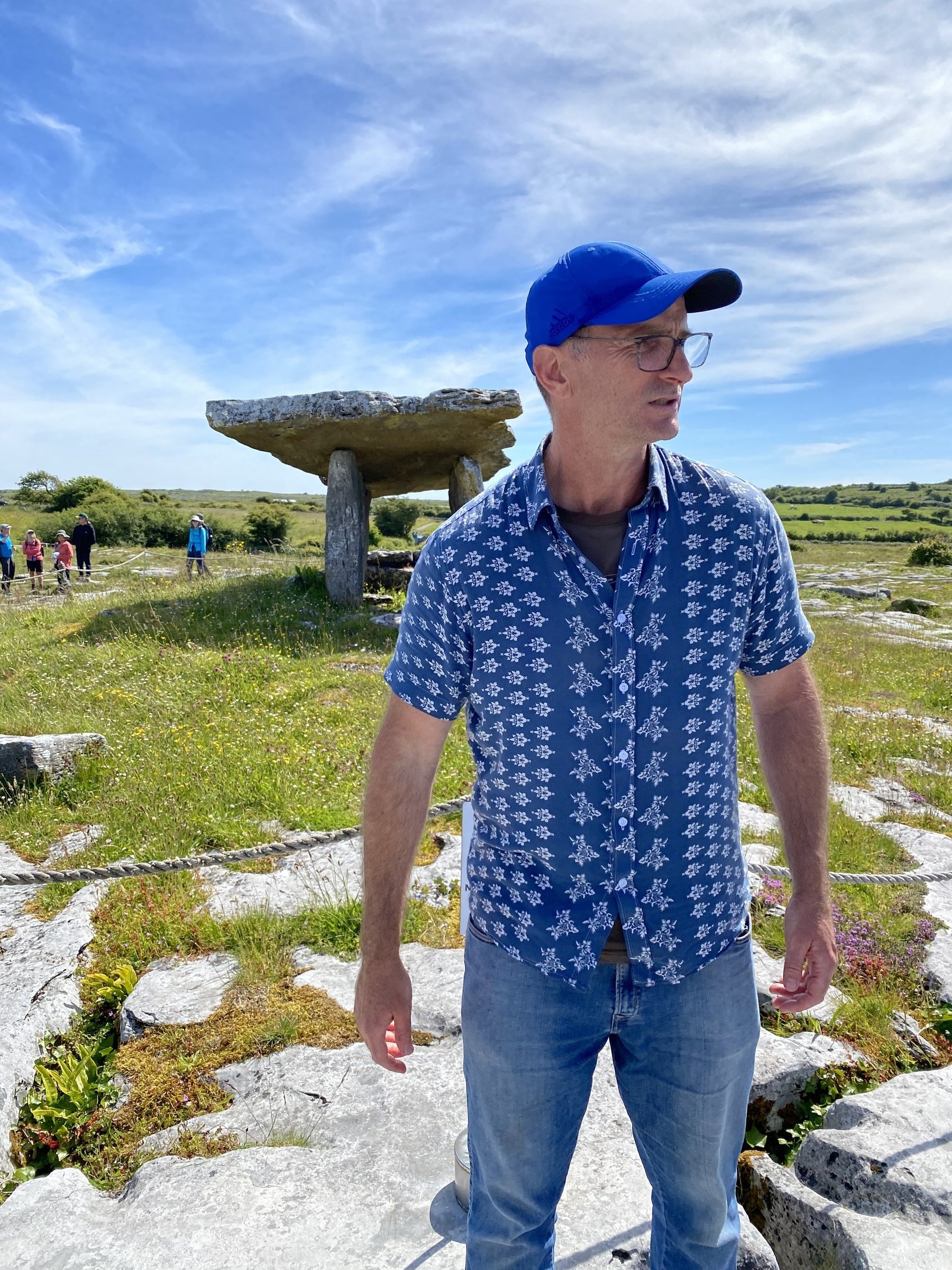 A man in a blue patterned shirt stands on limestone pavement with the ancient Poulnabrone dolmen rising behind him.