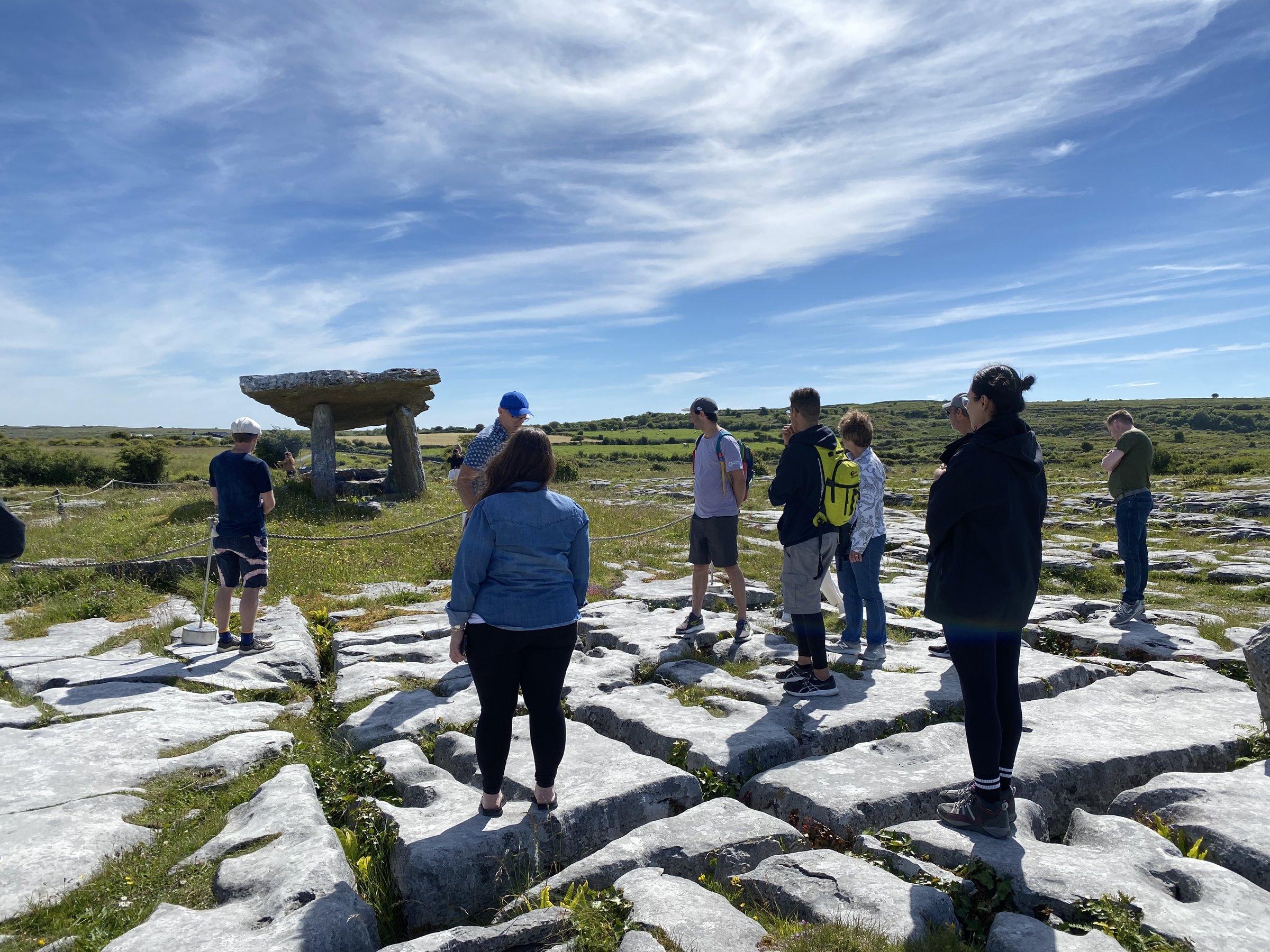 A group of visitors walks across the Burren's limestone pavement toward the Poulnabrone dolmen under a wide blue sky.