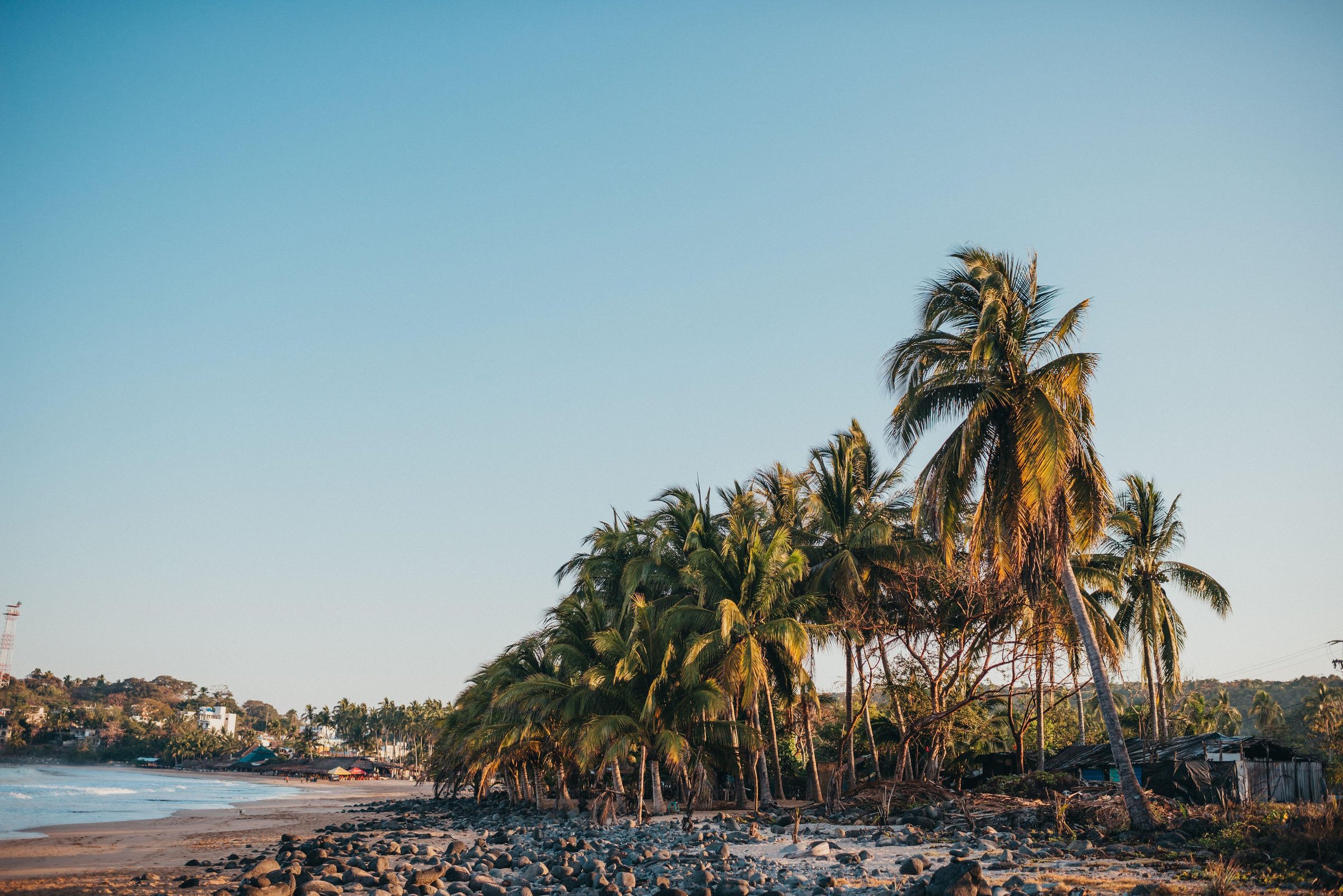 Palm trees and dark volcanic rocks line a sandy beach at golden hour, with the Pacific Ocean and a distant coastal village in Chacala, Mexico.