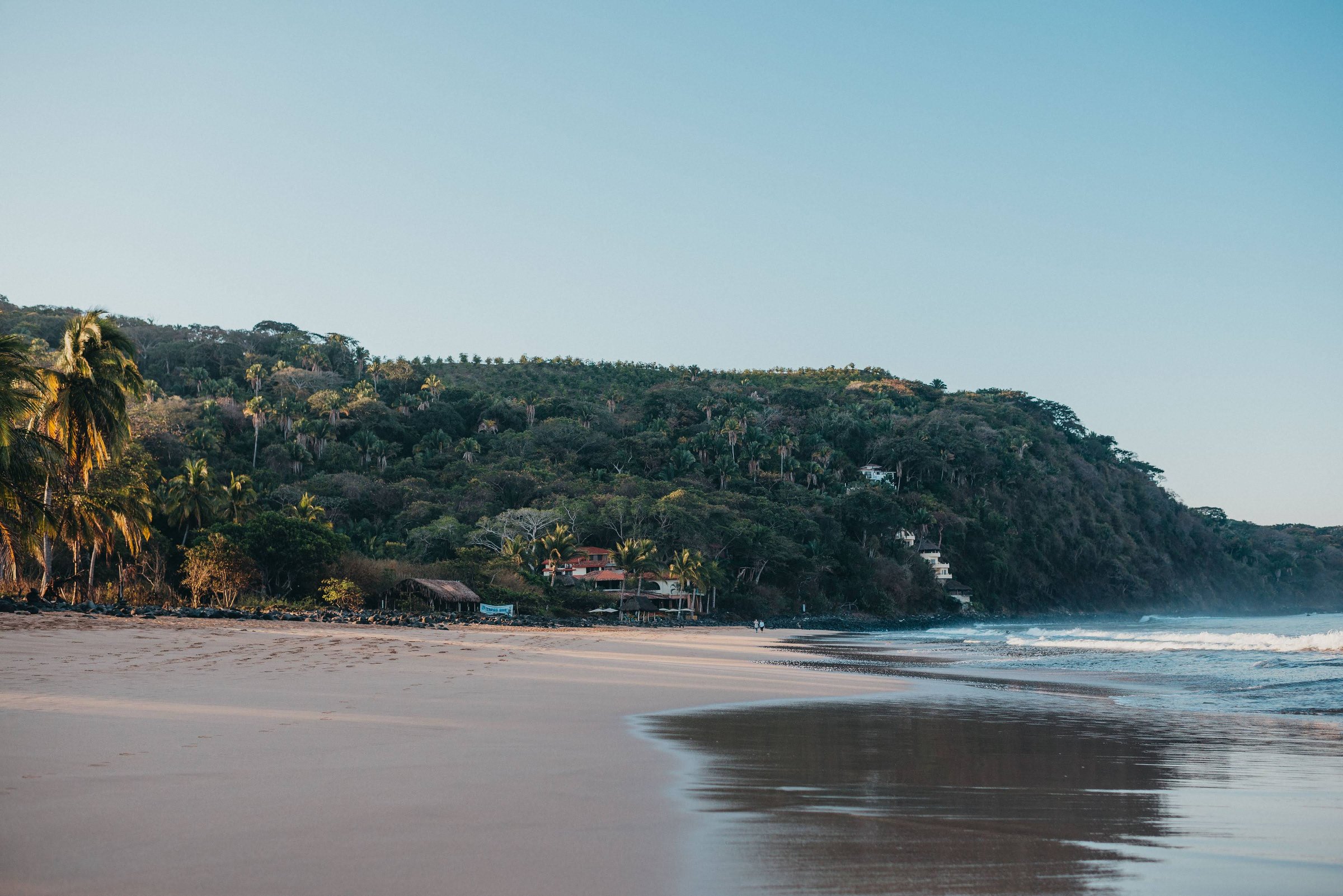 A wide crescent of sandy beach meets gentle surf at the base of a lush, palm-covered hillside in Chacala on Mexico's Pacific coast.