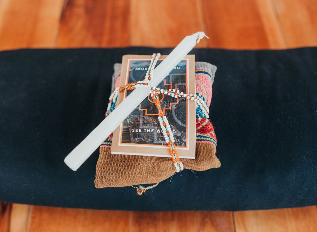 A small gift bundle with a white candle, Journey Peru notebook, and traditional Andean textile rests on a meditation cushion at a Sacred Valley retreat.