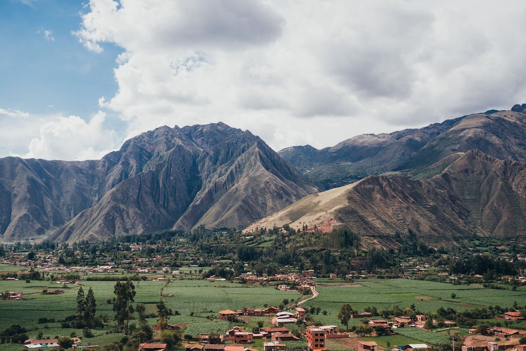 A wide view of a lush green village and terraced fields nestled at the base of towering Andes mountains in Peru's Sacred Valley.