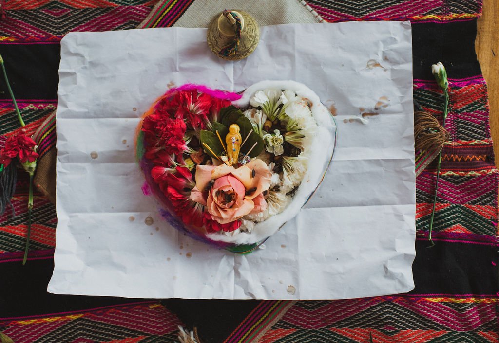 A heart-shaped ceremonial offering of red and white carnations and a small figurine rests on white paper over a vibrant Andean textile in the Sacred Valley.
