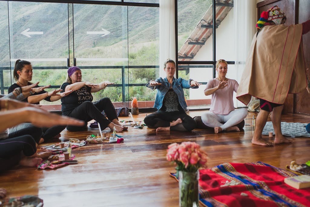 Retreat participants sit on a wooden floor with arms extended during a traditional ceremony led by a local practitioner, with green Andes slopes visible through floor-to-ceiling windows in the Sacred Valley.