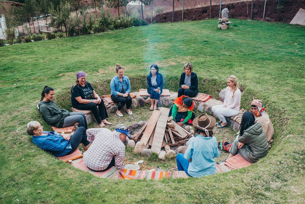 A group of retreat participants sits in a circle around a stone fire pit on a lush green lawn in the Sacred Valley, Peru.