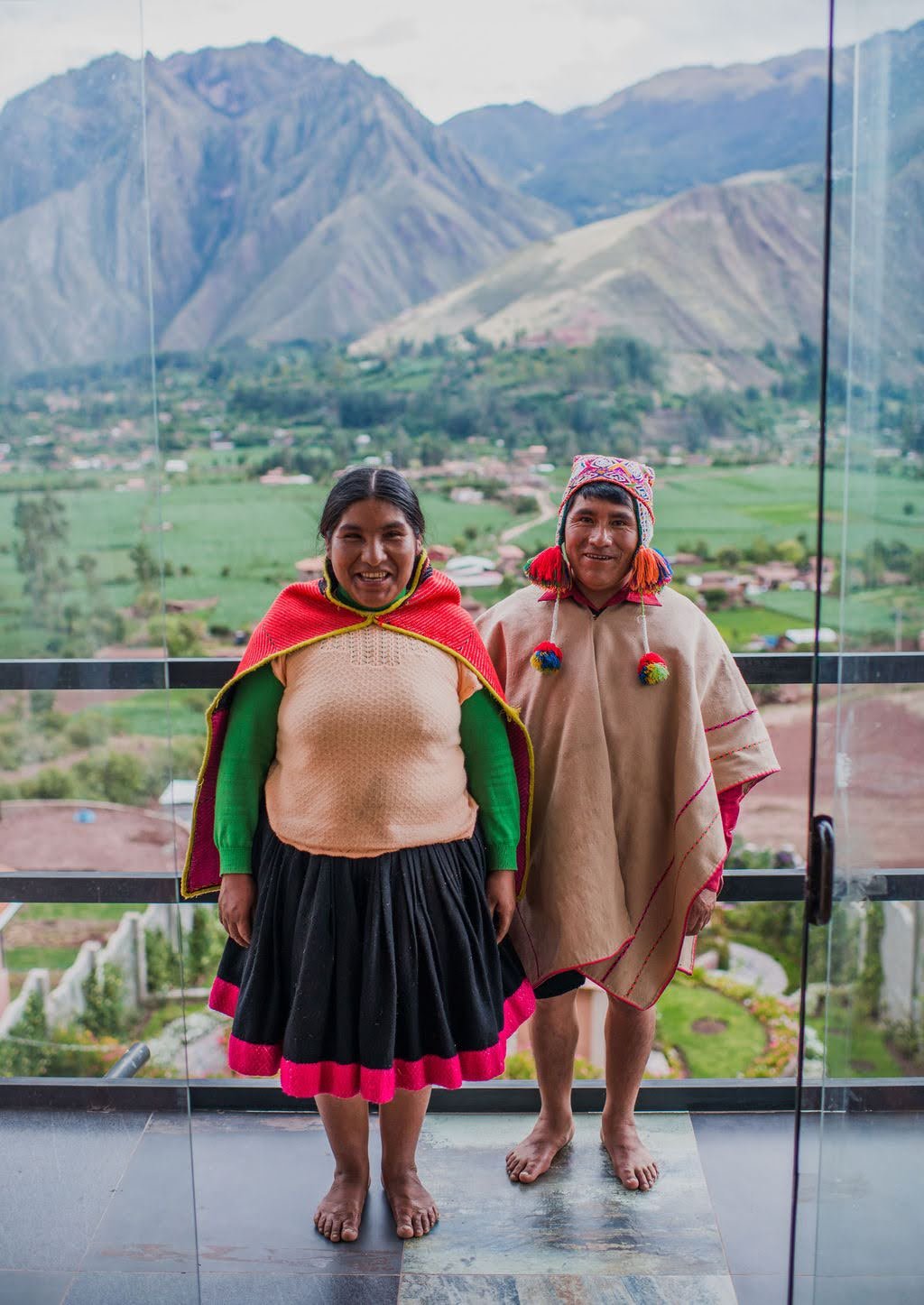 A smiling man and woman in traditional Andean clothing stand barefoot on a balcony overlooking the lush Sacred Valley and the Andes mountains.