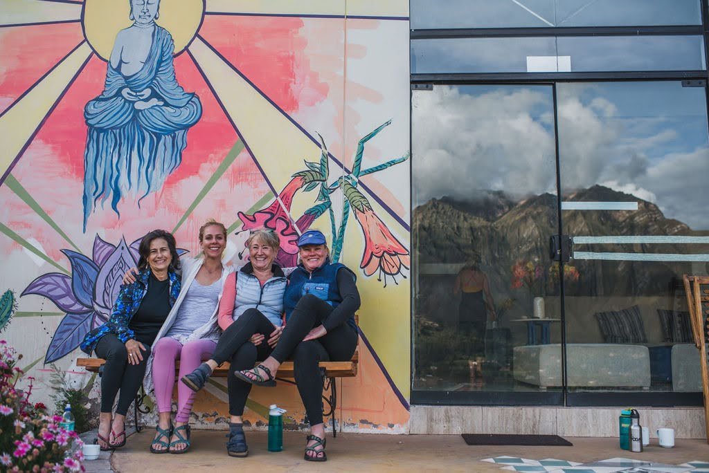 Four women smile while sitting on a bench in front of a colorful Buddha mural at a Sacred Valley retreat center, with the Andes reflected in a nearby glass door.