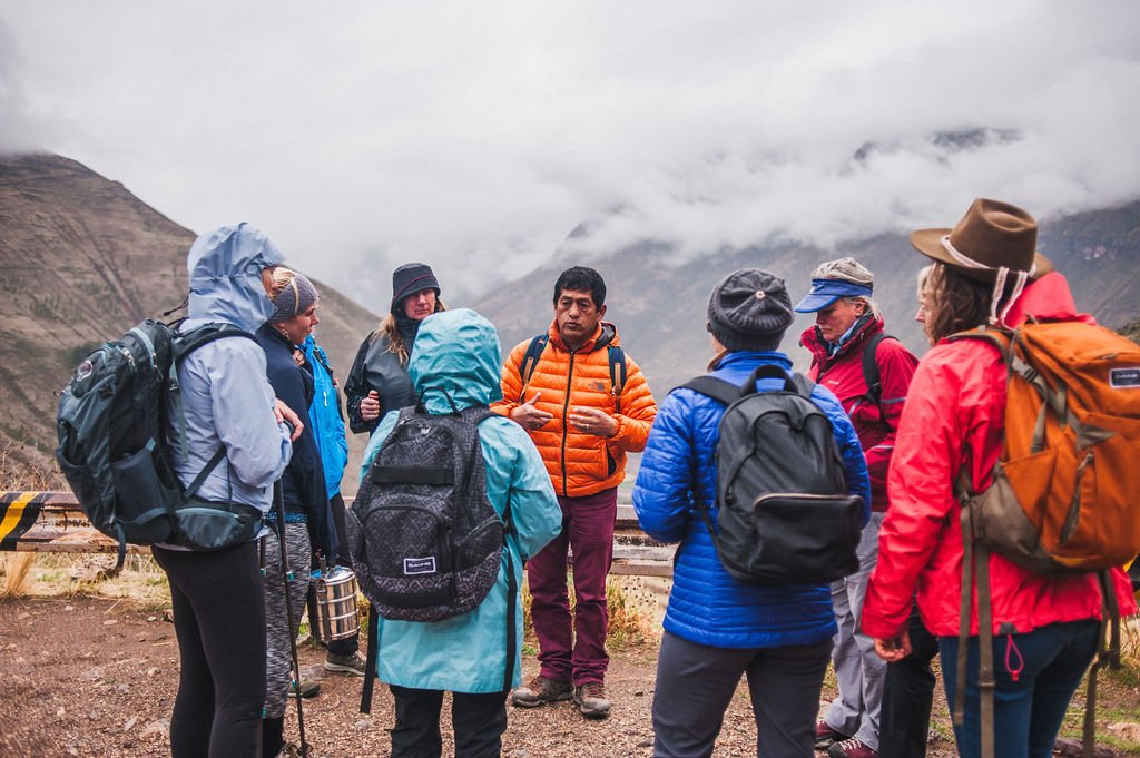A group of hikers in colorful jackets listens to a local guide in an orange puffer jacket on a misty mountain trail in the Sacred Valley.