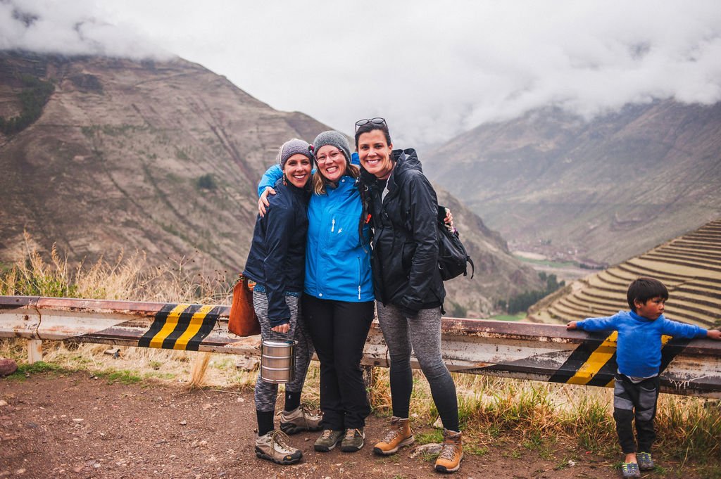 Three women hug and smile on a roadside overlooking misty Andes mountains and ancient agricultural terraces in the Sacred Valley.