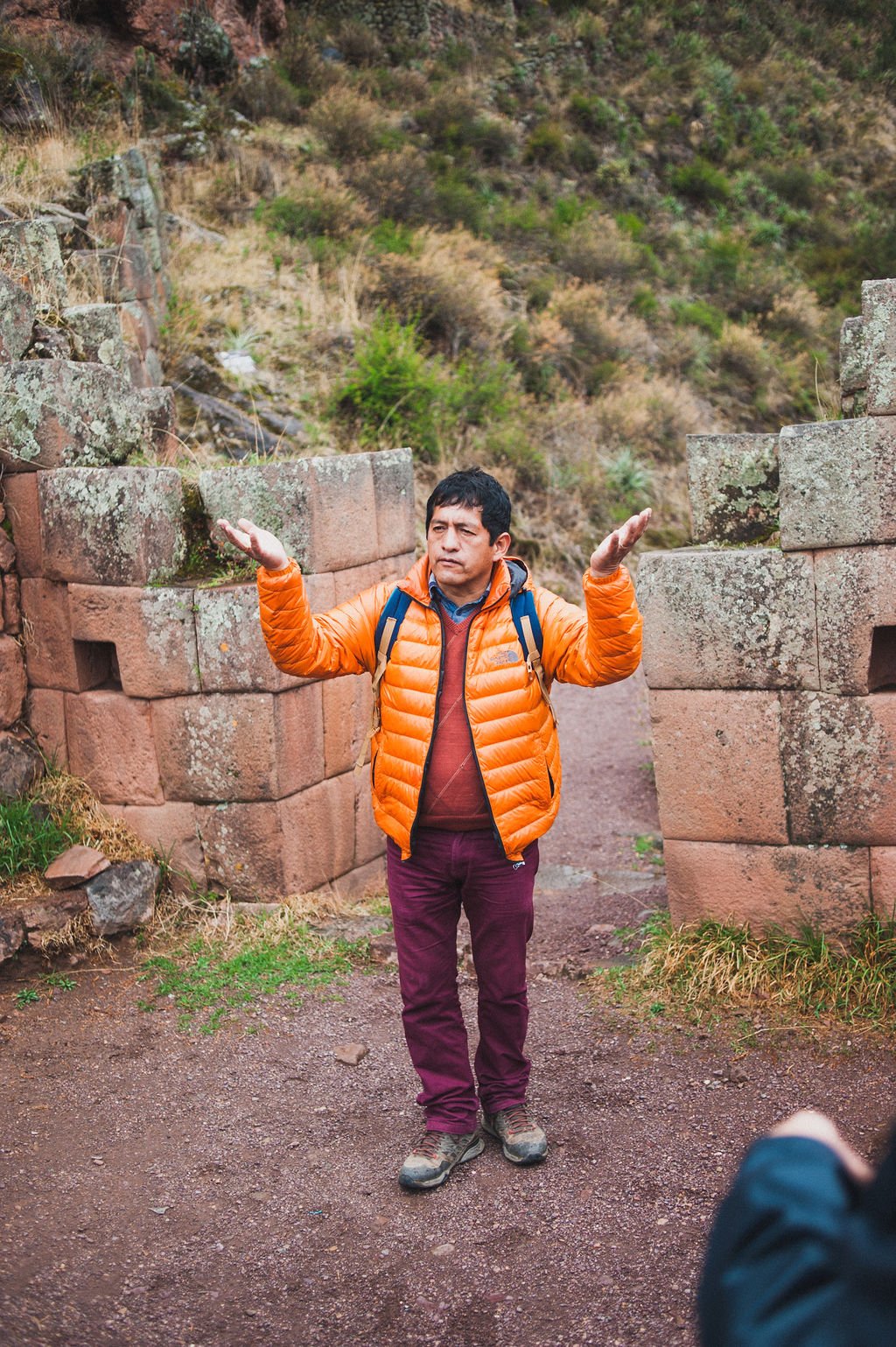 A guide in a bright orange jacket gestures while standing between ancient Inca stone walls on a hillside in the Sacred Valley.