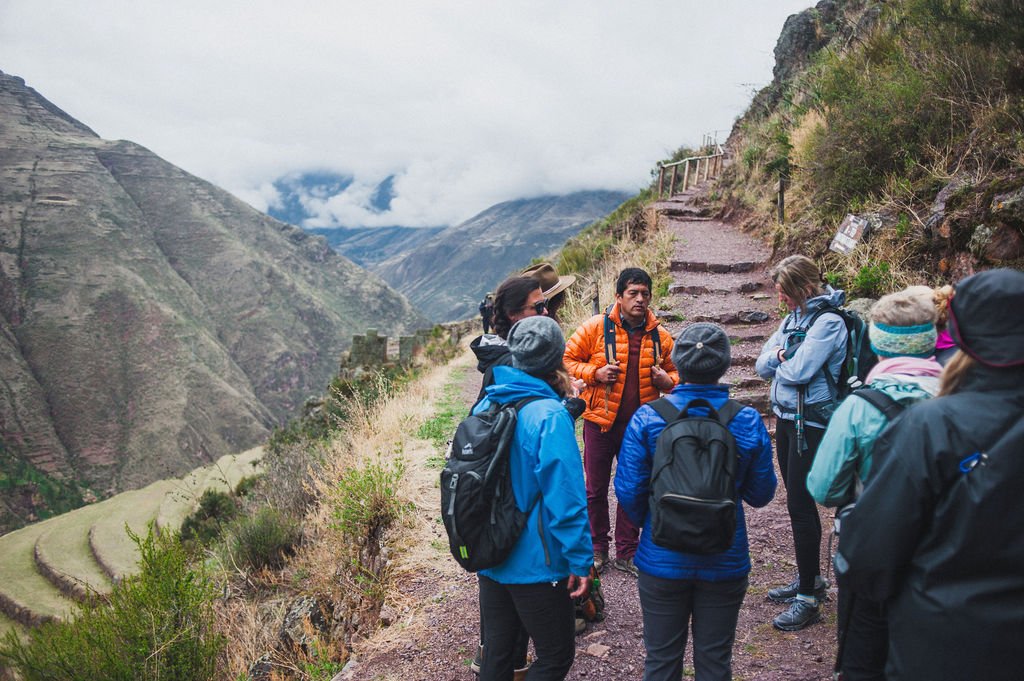 A group of travelers listens to their guide on a rugged mountain path overlooking the ancient stone terraces of Pisac in the Sacred Valley.