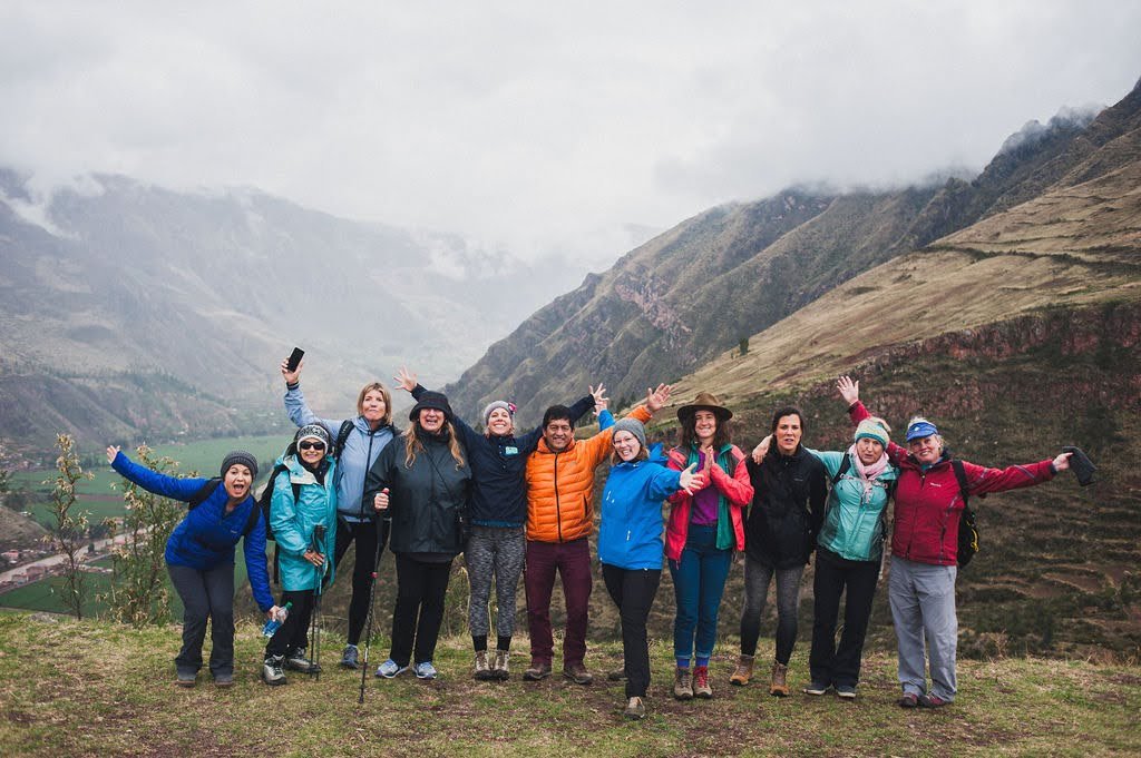 A joyful group of eleven hikers raises their arms on a grassy ridge overlooking the misty Andes and the green floor of the Sacred Valley.
