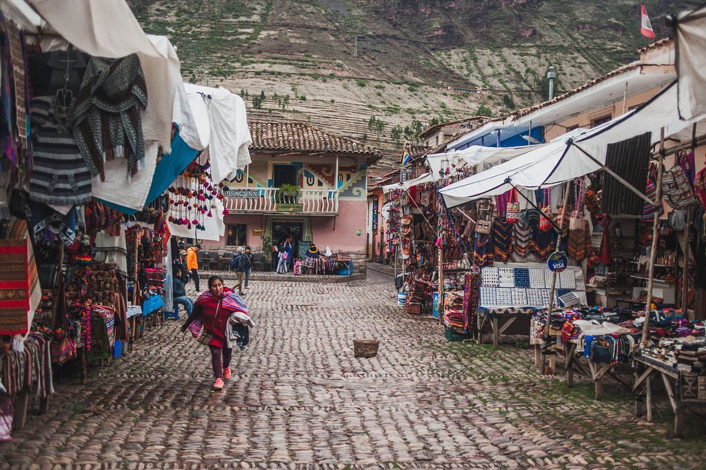 A woman in a red jacket walks across a cobblestone street lined with colorful market stalls selling traditional textiles in Pisac.