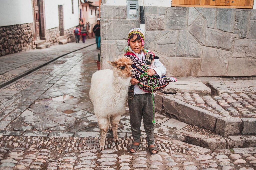 A young boy in traditional Andean dress stands on a wet cobblestone street in Cusco, holding a small lamb in a woven sling beside a white alpaca.