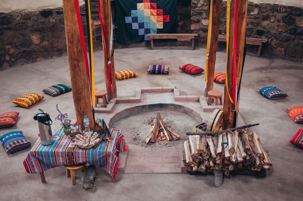 A circular ceremonial room in the Sacred Valley features a central fire pit surrounded by colorful woven cushions, traditional textiles, and a Chakana flag on a stone wall.