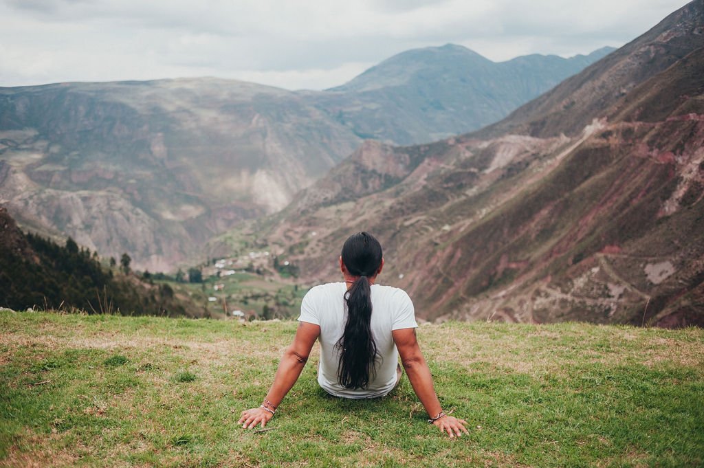 A person with long dark hair sits on a grassy ridge looking out over a vast, cloud-covered mountain valley in the Sacred Valley.